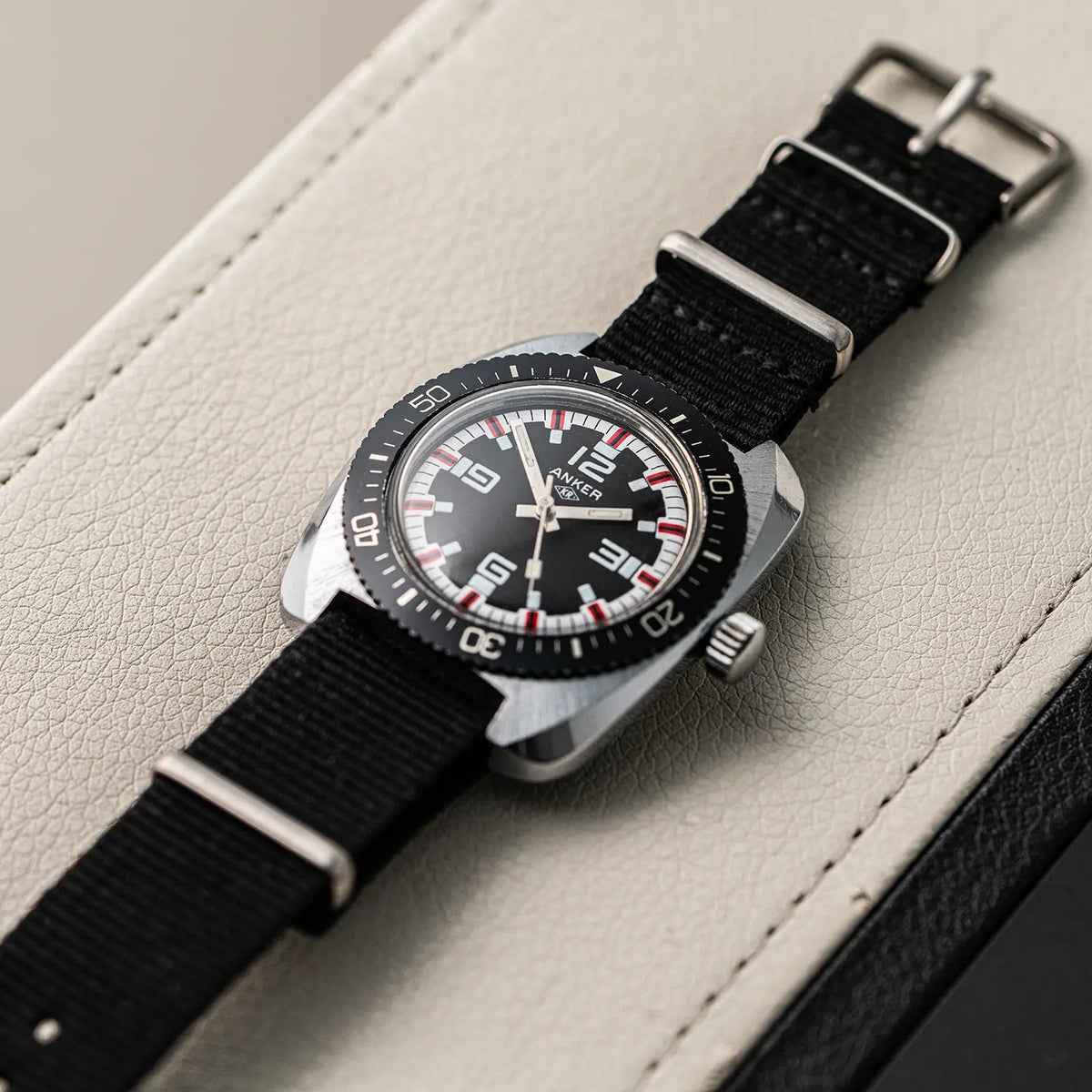 A vintage-style wristwatch Anker KR Diver, Cal. UMF 24, Men’s Diving Vintage Watch with a black dial, silver-toned metal case, and black fabric strap, photographed diagonally on a light beige textured leather surface.