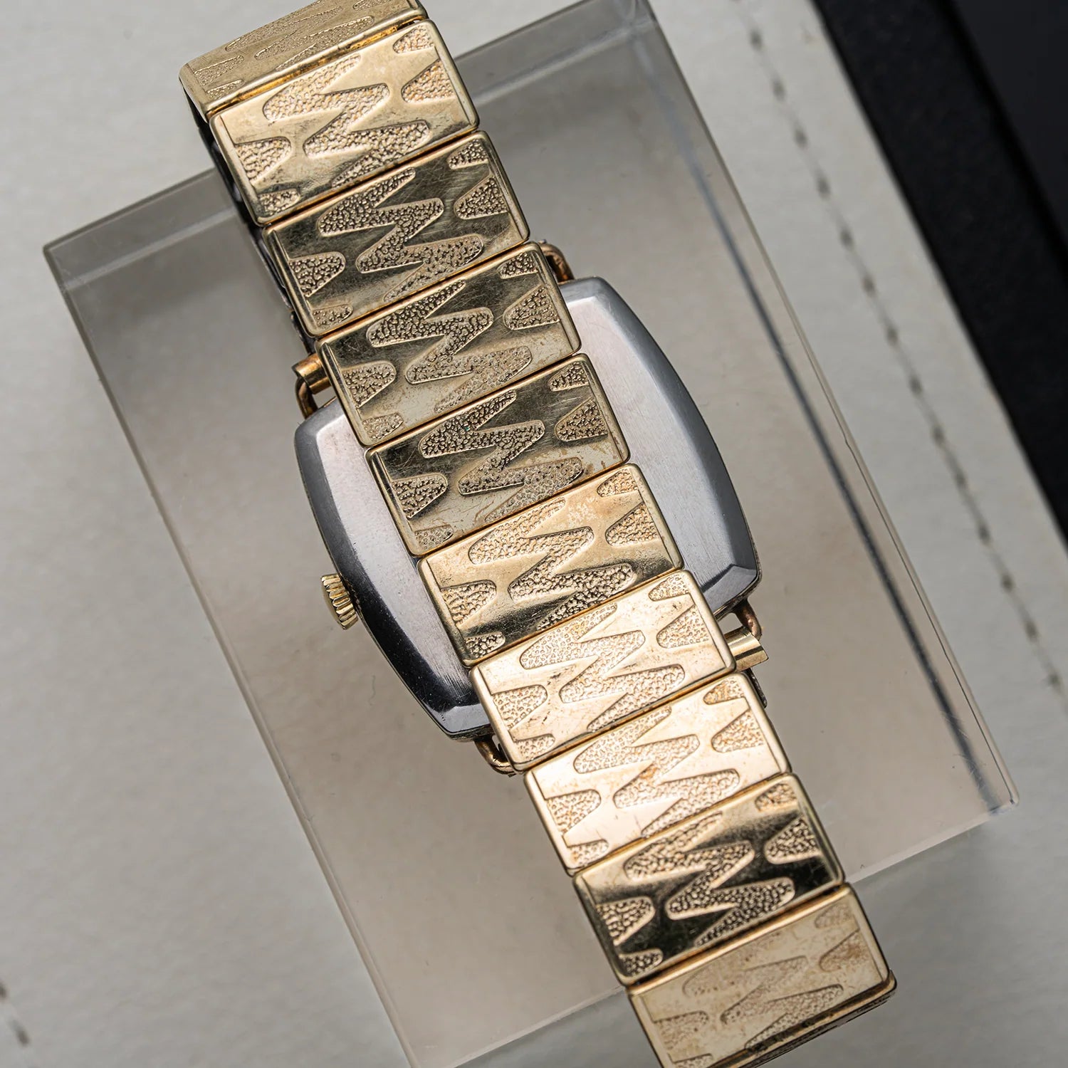 A close-up of a vintage gold-tone watch Bergana PUW 460 with a distinctive textured link bracelet, resting on a clear display stand.