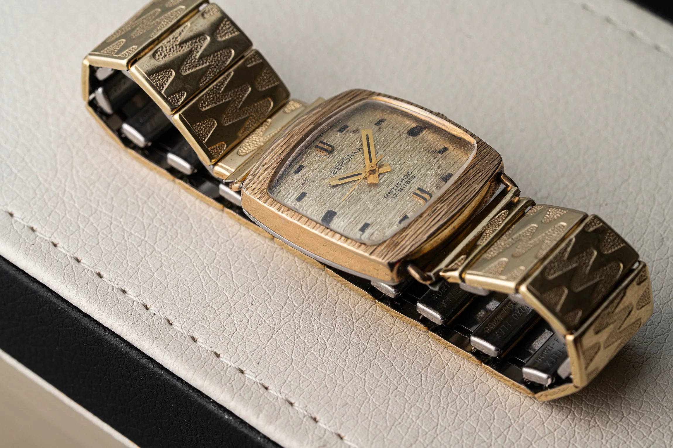 A vintage gold-tone wristwatch Bergana PUW 460 with a textured chevron-patterned metal bracelet and a square dial, resting on a light beige textured leather surface.