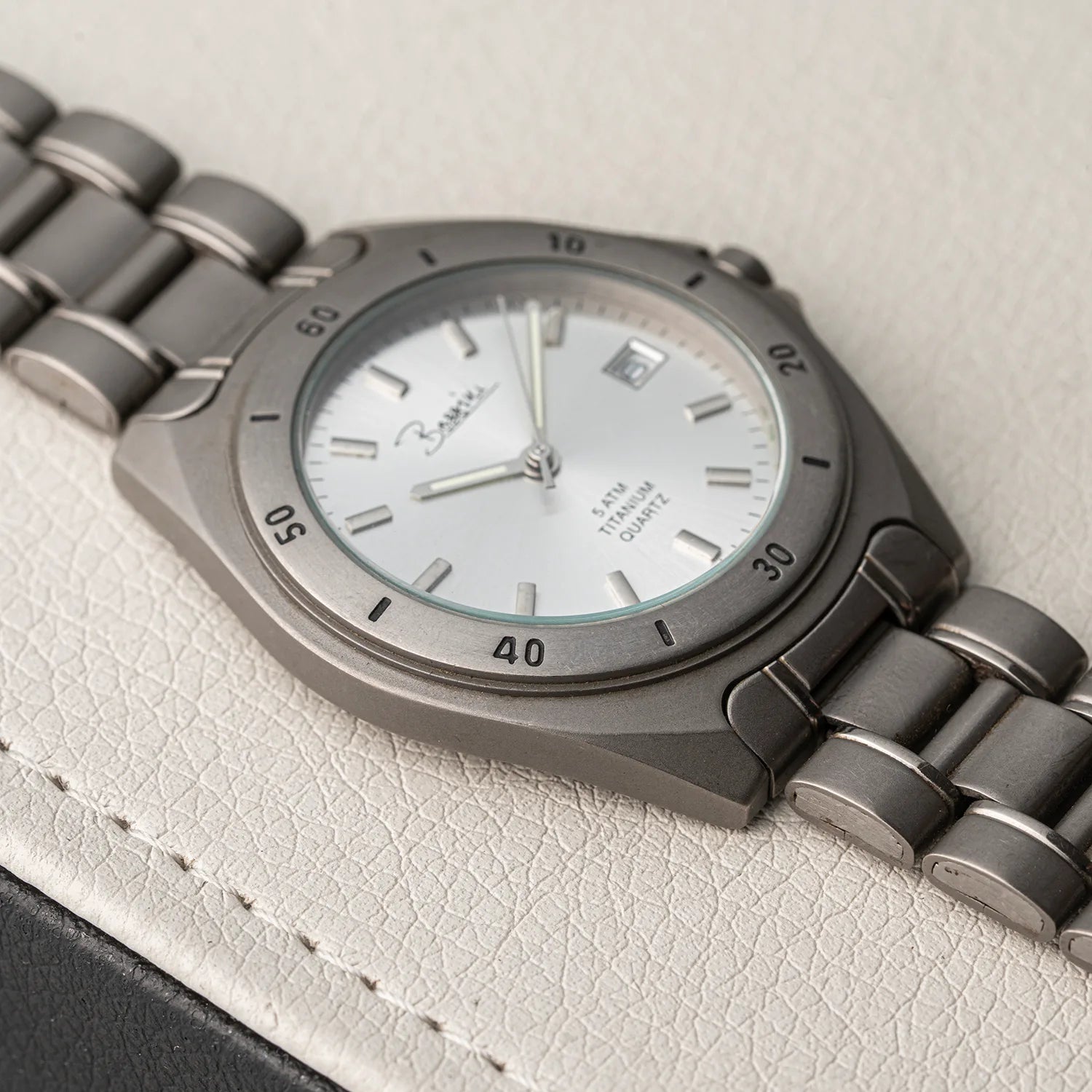 A close-up of a wristwatch Bossini Titanium with a matte gray titanium bracelet and a white dial, resting on a textured off-white leather surface.