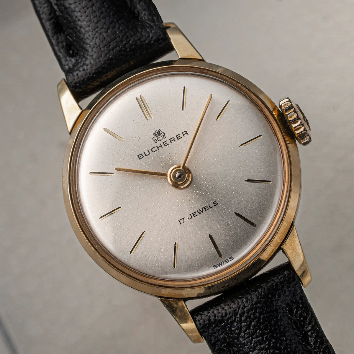 A vintage-style wristwatch Bucherer, Ref. 16.238, Women’s Manual Wind with a round silver dial, gold-toned case, black textured leather strap, and slim stick indices, photographed close up on a neutral gray background.