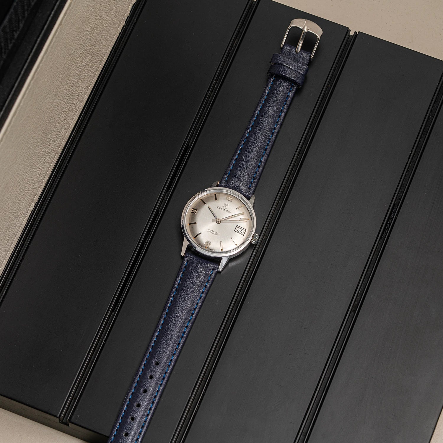 A vintage-style wristwatch Delbana, Cal. AS 1951 with a silver-toned case, white dial, and blue leather strap, laid diagonally on a dark textured surface.
