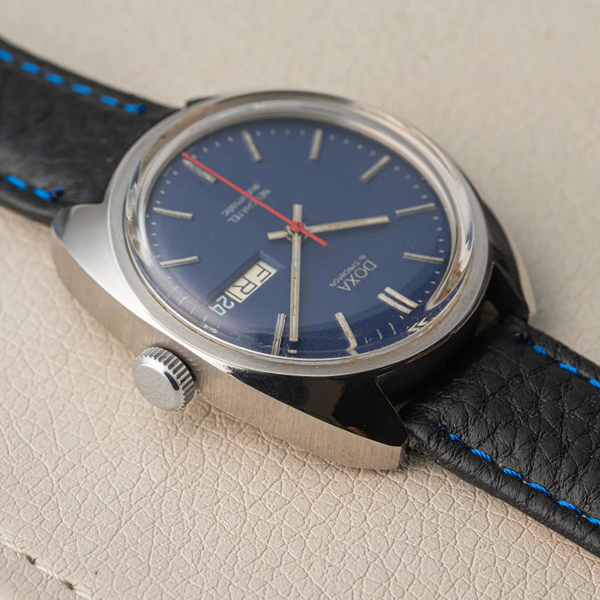 A close-up of a Doxa Neuchatel, Ref. 565.005.56, Cal. AS 5206, Men’s Automatic wristwatch with a blue dial, silver-tone case, and black leather strap with blue stitching, resting on a textured beige surface.