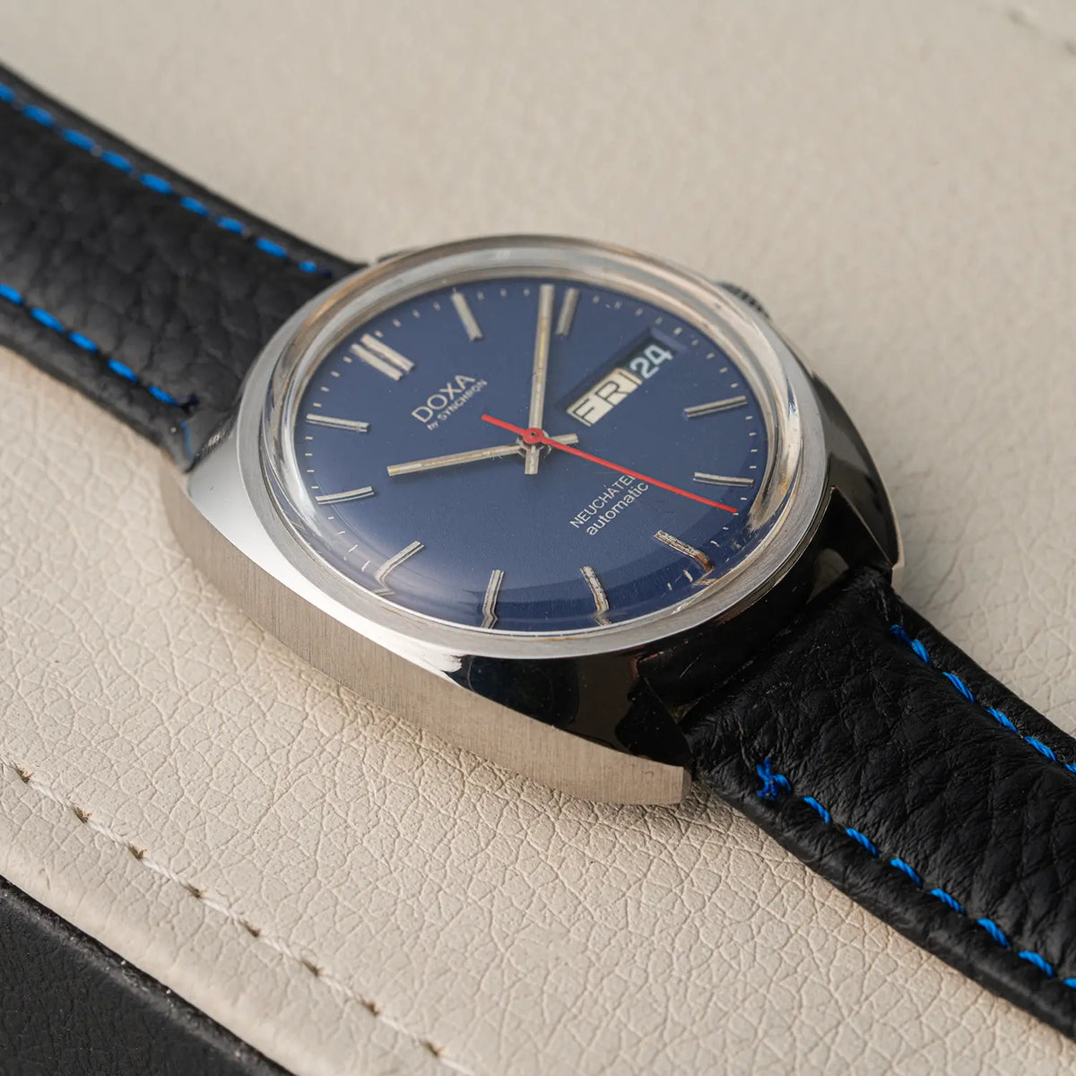 A close-up of a Doxa Neuchatel, Ref. 565.005.56, Cal. AS 5206, Men’s Automaticwristwatch with a blue dial, silver-toned case, and black leather strap with bright blue stitching, resting on a textured beige surface.