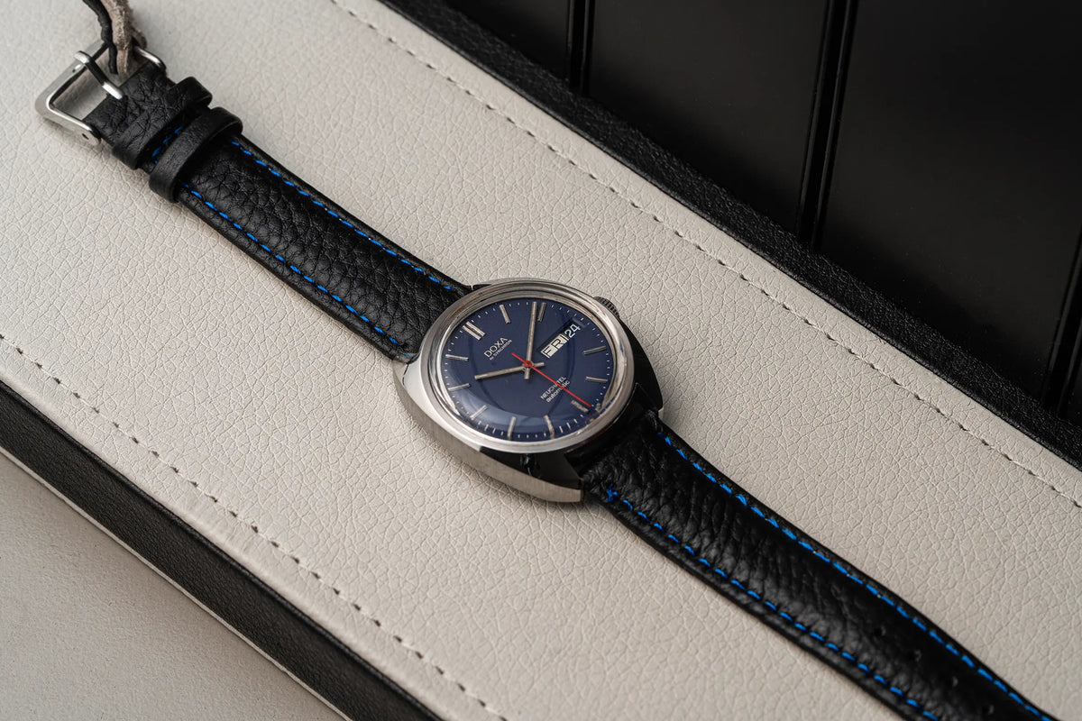 A close-up of a wristwatch Doxa Neuchatel, Ref. 565.005.56, Cal. AS 5206, Men’s Automatic with a silver-toned case, blue dial, and black textured leather strap with bright blue contrast stitching, resting on a light cream textured surface.