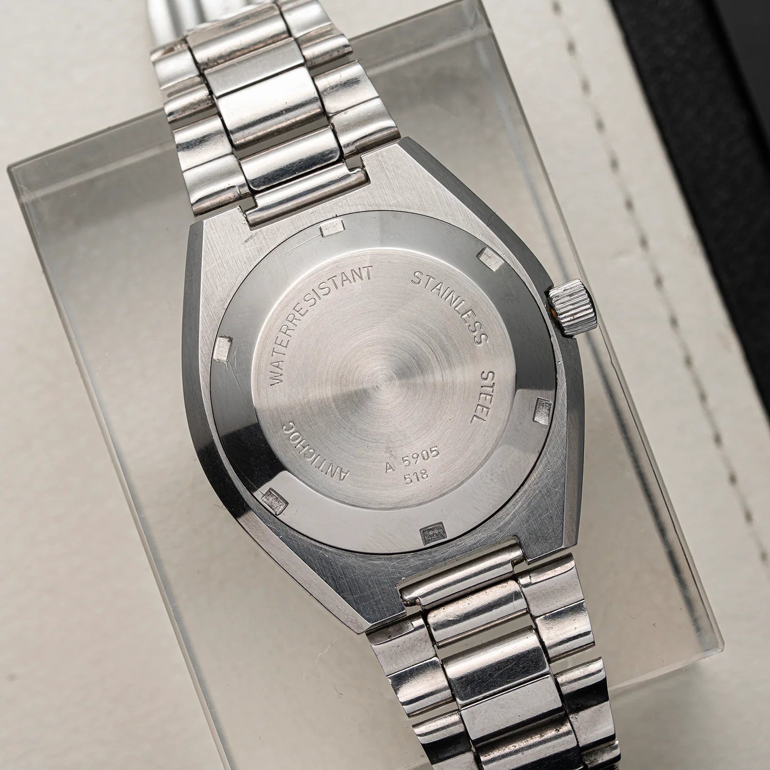 Close-up of the back of a stainless steel wristwatch Dugena Tropica with a metal link bracelet, resting on a clear display stand. The case is brushed silver with engraved text including 'WATERRESISTANT', 'STAINLESS STEEL', and 'Oyster'.