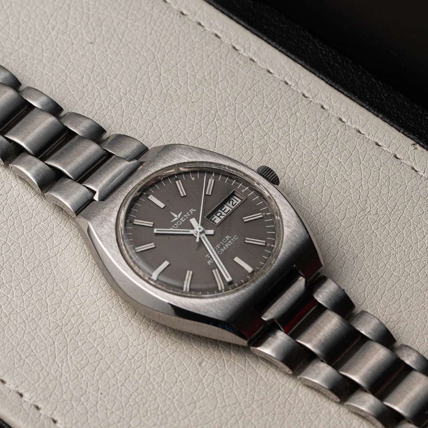 A vintage-style wristwatch Dugena Tropica with a brushed silver-tone metal case and multi-link bracelet, featuring a dark gray dial with silver indices and a day/date window. The watch is photographed diagonally on a textured light beige leather surface.