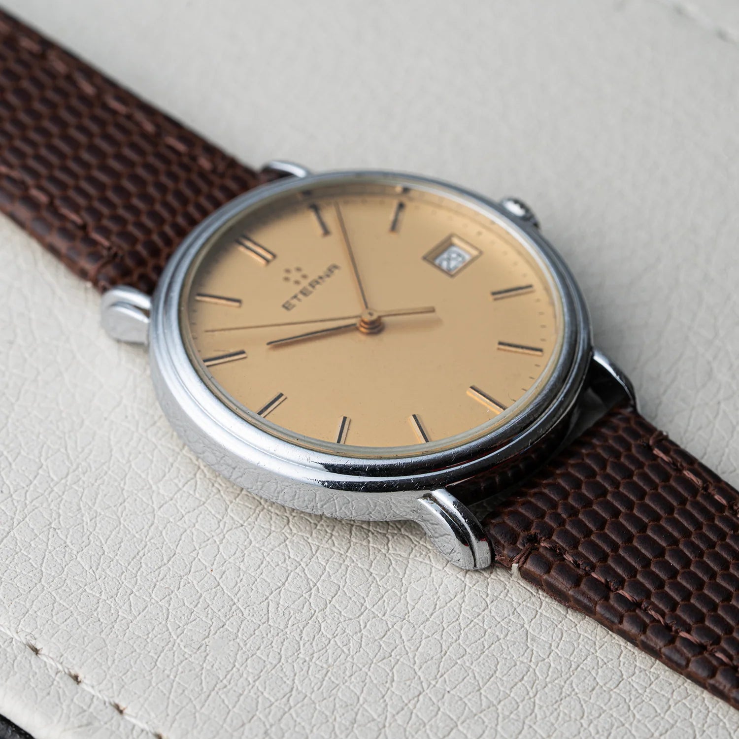A vintage-style wristwatch Eterna Quartz, Cal. ETA 955.112 with a silver-toned oval case, a warm beige dial, and a dark brown textured leather strap, photographed on a light cream textured surface.