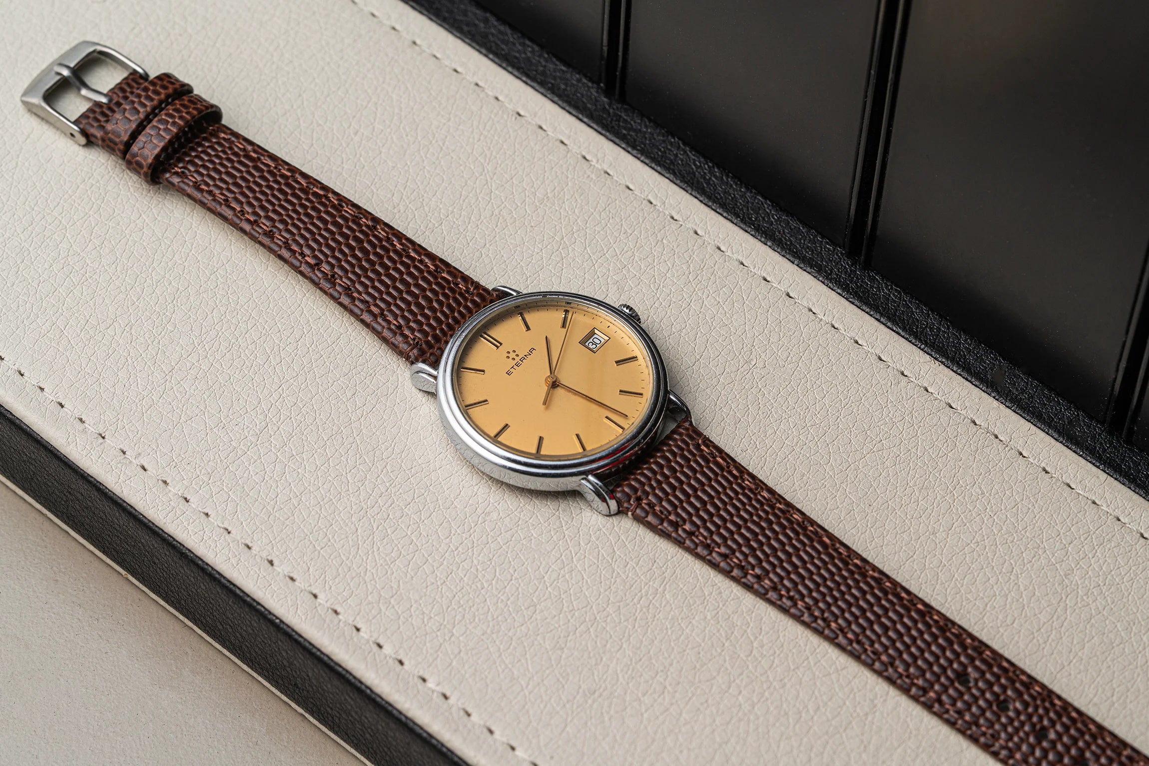 A vintage-style wristwatch Eterna Quartz, Cal. ETA 955.112 with a brown textured leather strap and a round silver-tone case, resting diagonally on a cream-colored textured surface.