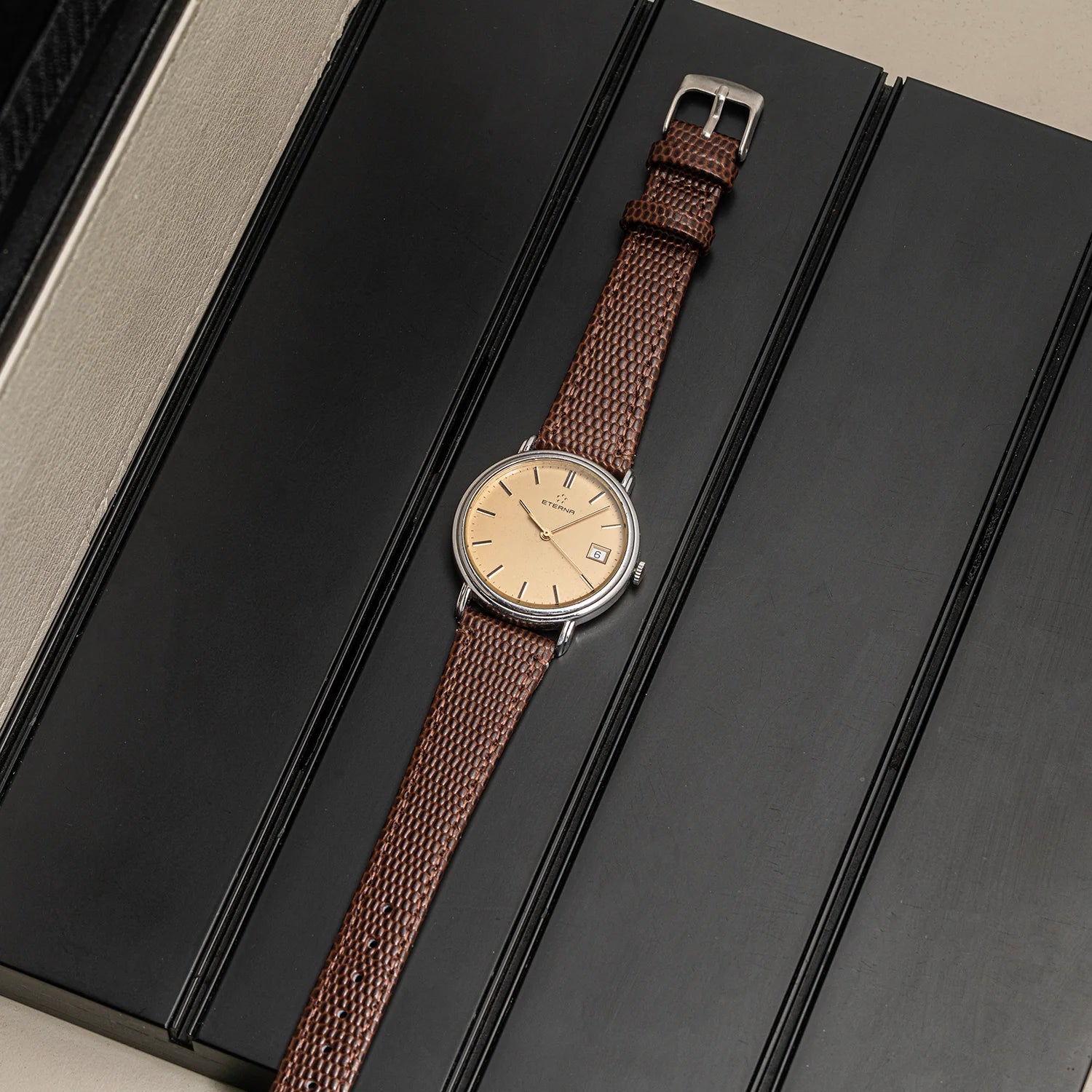 A vintage-style wristwatch Eterna Quartz, Cal. ETA 955.112 with a brown woven strap and a beige dial, displayed diagonally on a black presentation box.