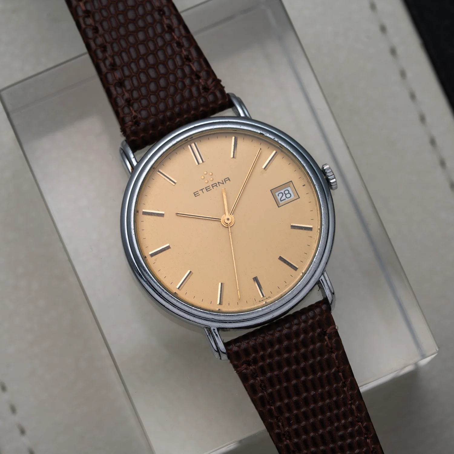 A vintage-style wristwatch Eterna Quartz, Cal. ETA 955.112 with a round silver-toned case, beige dial, and dark brown textured strap, resting on a light gray presentation box. The brand name 'ETERN' is visible on the dial.