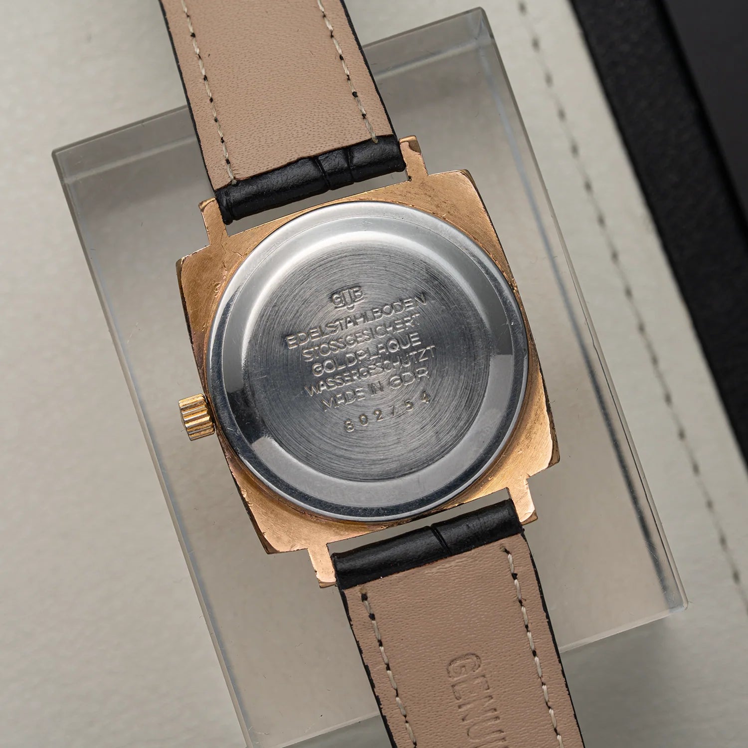Close-up of the back of a vintage wristwatch Glashutte Bison with a gold-tone case, a silver-toned metal case back engraved with text, and a beige leather strap with black stitching, displayed on a clear acrylic stand.