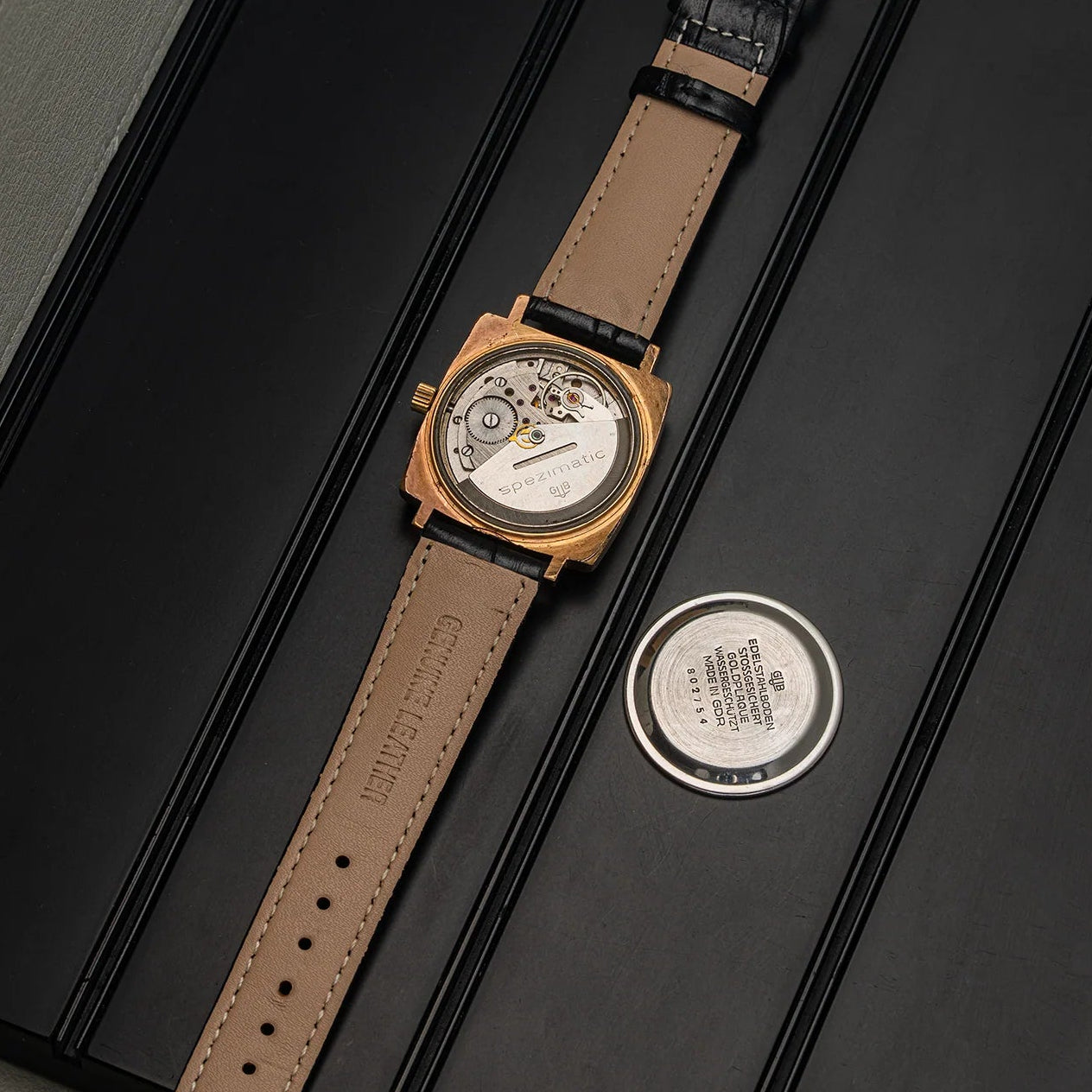 A luxury watch Glashutte Bison with a gold-toned case, a tan leather strap, and a visible mechanical movement, displayed diagonally on a black presentation box next to its circular metal backplate.