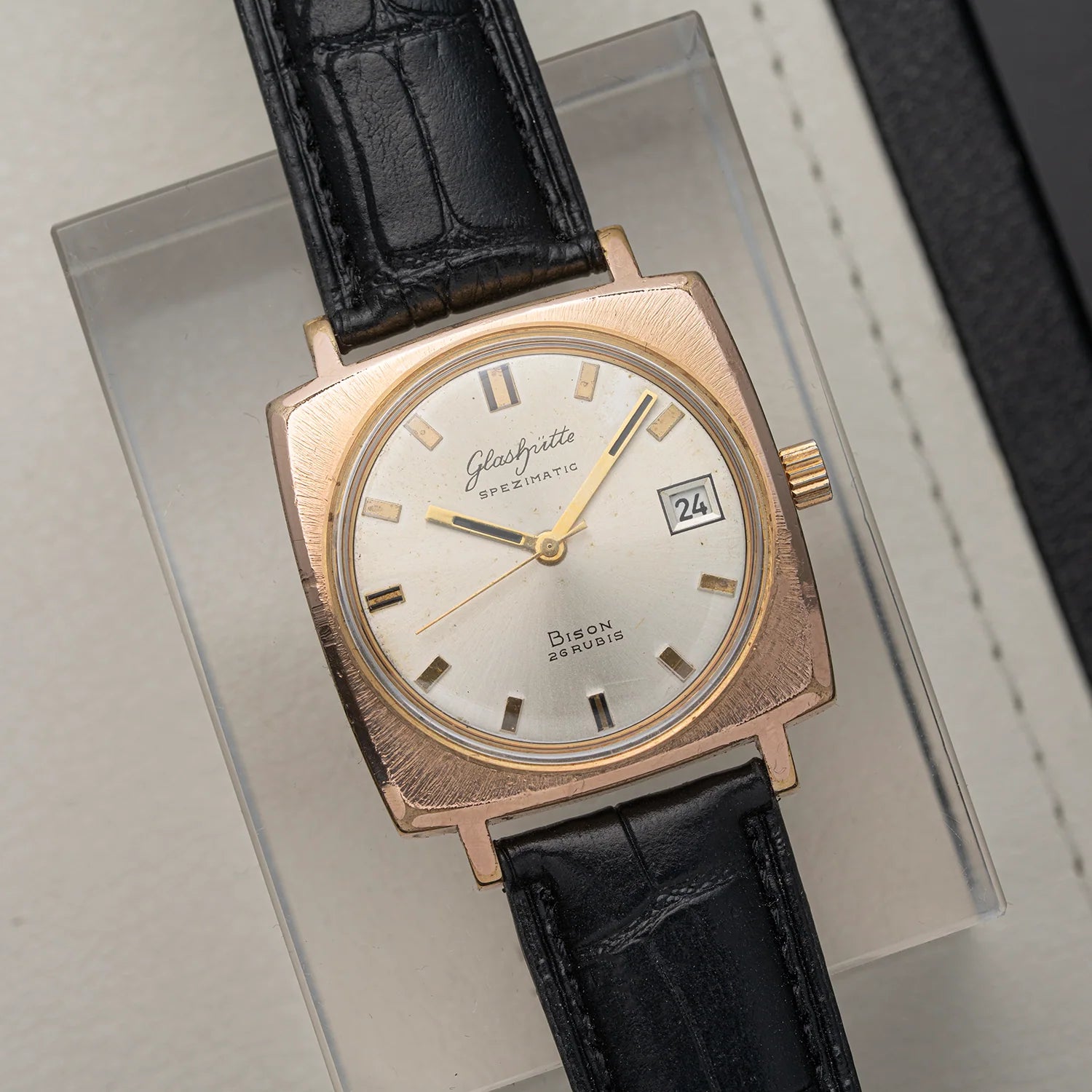 A vintage Glashutte Bison automatic watch with a rose gold-toned square case, white dial, and black leather strap, displayed on a clear acrylic stand.