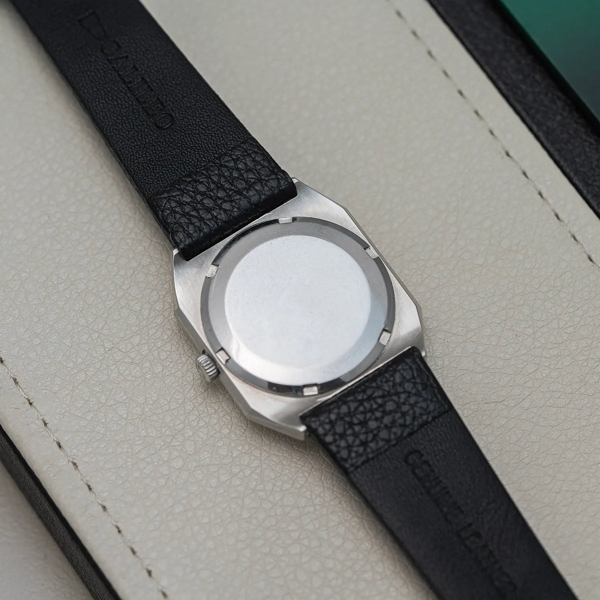 Close-up of a wristwatch Longines Octagon, Ref. 4817-4, Cal. L633.1, Men’s Automatic, 1976 with a silver-toned metal case and a black textured leather strap, laid diagonally on a light beige textured surface.