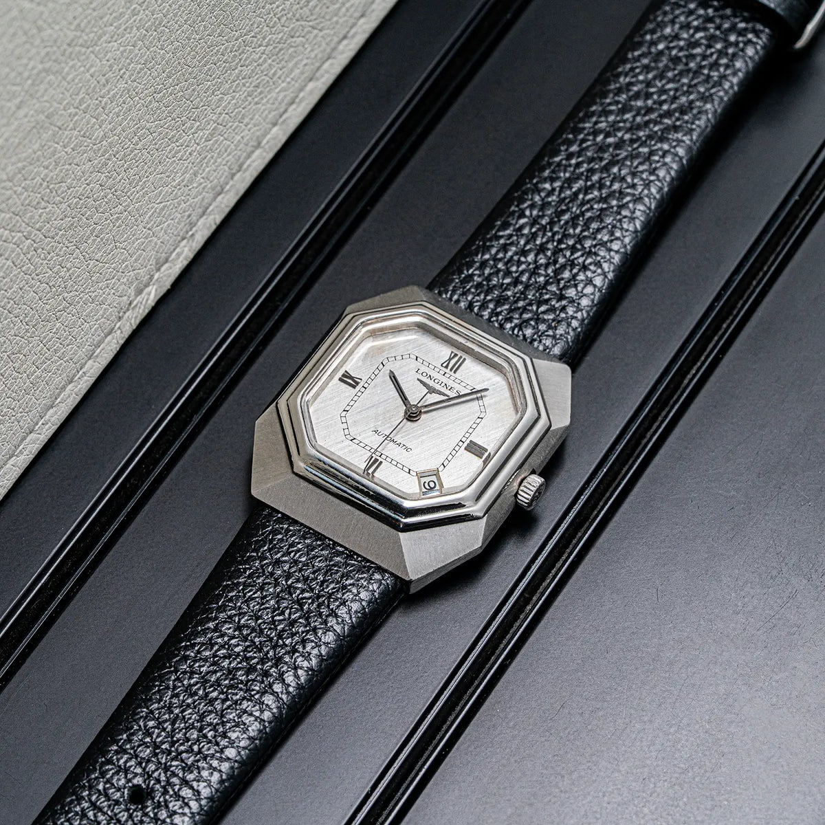 A close-up of a wristwatch Longines Octagon, Ref. 4817-4, Cal. L633.1, Men’s Automatic, 1976 with an octagonal silver-toned case, white dial, and textured black leather strap, resting on a dark surface next to a light textured panel.