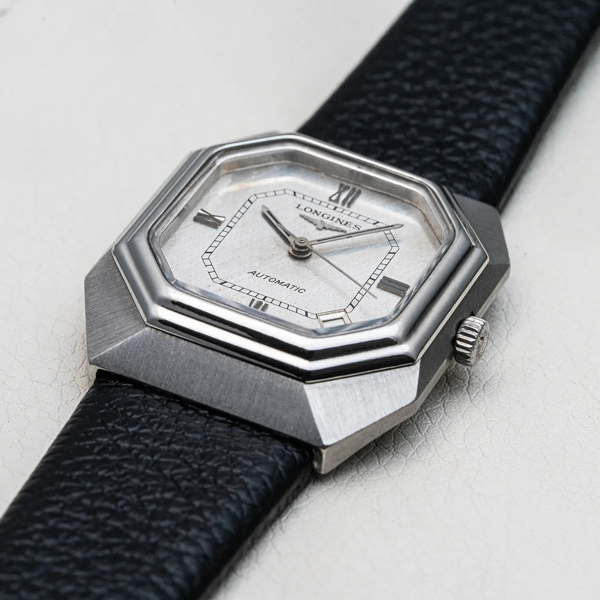 A close-up shot of a Longines Automatic watch with an octagonal silver-tone case, a white dial, and a textured black leather strap, set against a light gray textured background.