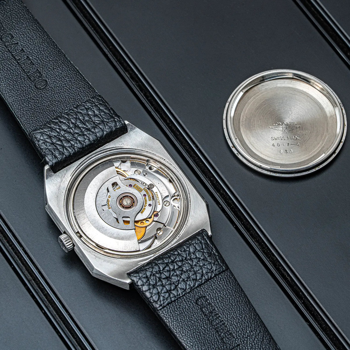 A close-up of a watch Longines Octagon, Ref. 4817-4, Cal. L633.1, Men’s Automatic, 1976 shown from the back, with a silver-toned case, black textured leather strap, and exposed mechanical movement visible. The back of the case is placed separately on a dark surface to the right.