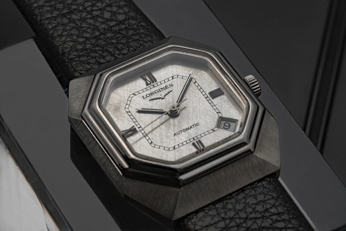 A close-up of a LLongines Octagon, Ref. 4817-4, Cal. L633.1, Men’s Automatic, 1976 octagonal automatic wristwatch with a textured dark gray metal case, white dial, and black textured strap, resting on a dark reflective surface.
