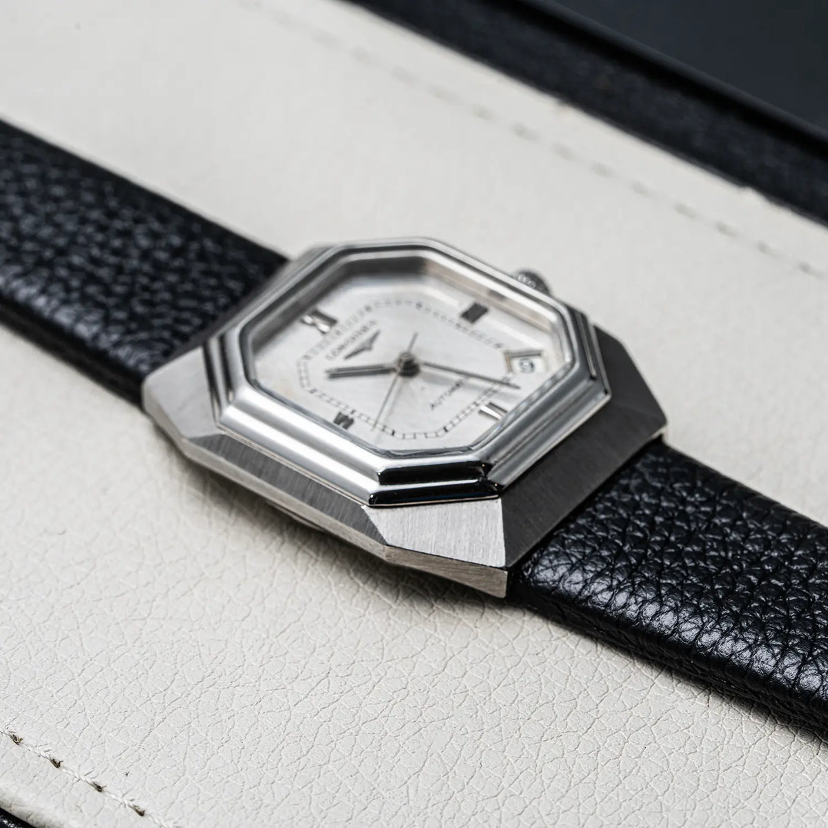 A close-up of a wristwatch Longines Octagon, Ref. 4817-4, Cal. L633.1, Men’s Automatic, 1976 with an angular silver-tone metal case and a white dial, mounted on a black textured leather strap against a light cream leather background.