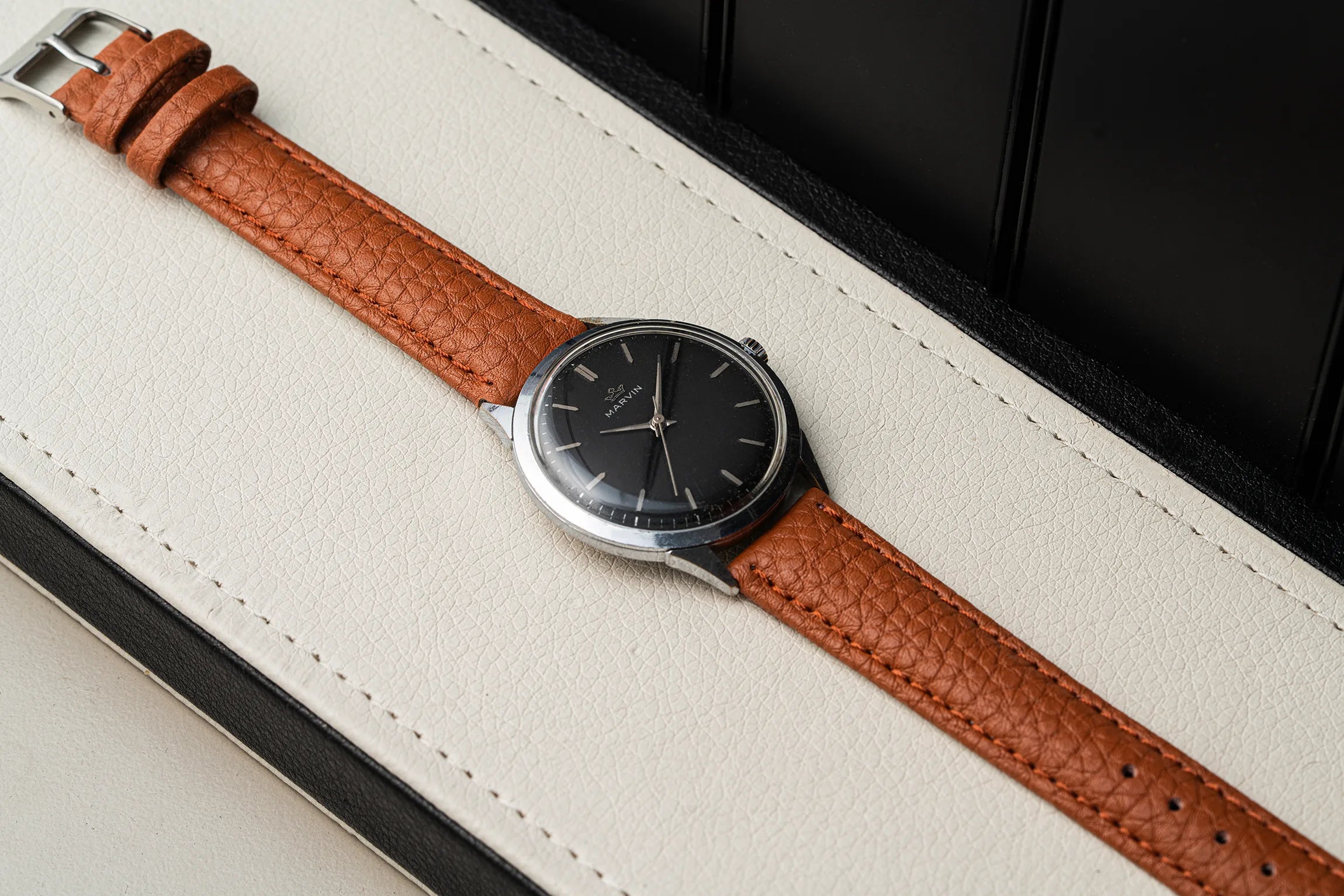 A vintage-style wristwatch Marvin Cal. 560 with a textured brown leather strap and a black dial, resting diagonally on a cream-colored leather surface with a black background.