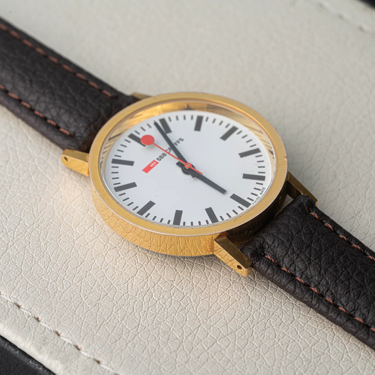 A close-up of a vintage-style Mondaine Official Swiss Railways Watch wristwatch with a gold case, white dial, black hour markers and hands, a red second hand and dot, and a dark brown leather strap with contrast stitching, resting on a textured off-white surface.