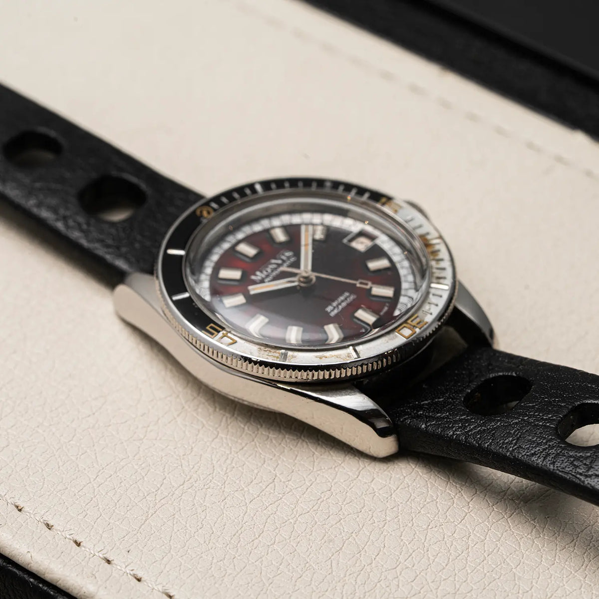 A vintage-style stainless steel watch Monvis Bakelite Diver, 20 ATM, Ref. 1.18, Cal. ETA 2783, Men’s Diving with a black dial, black bezel, and black leather strap, resting on a light beige textured surface.