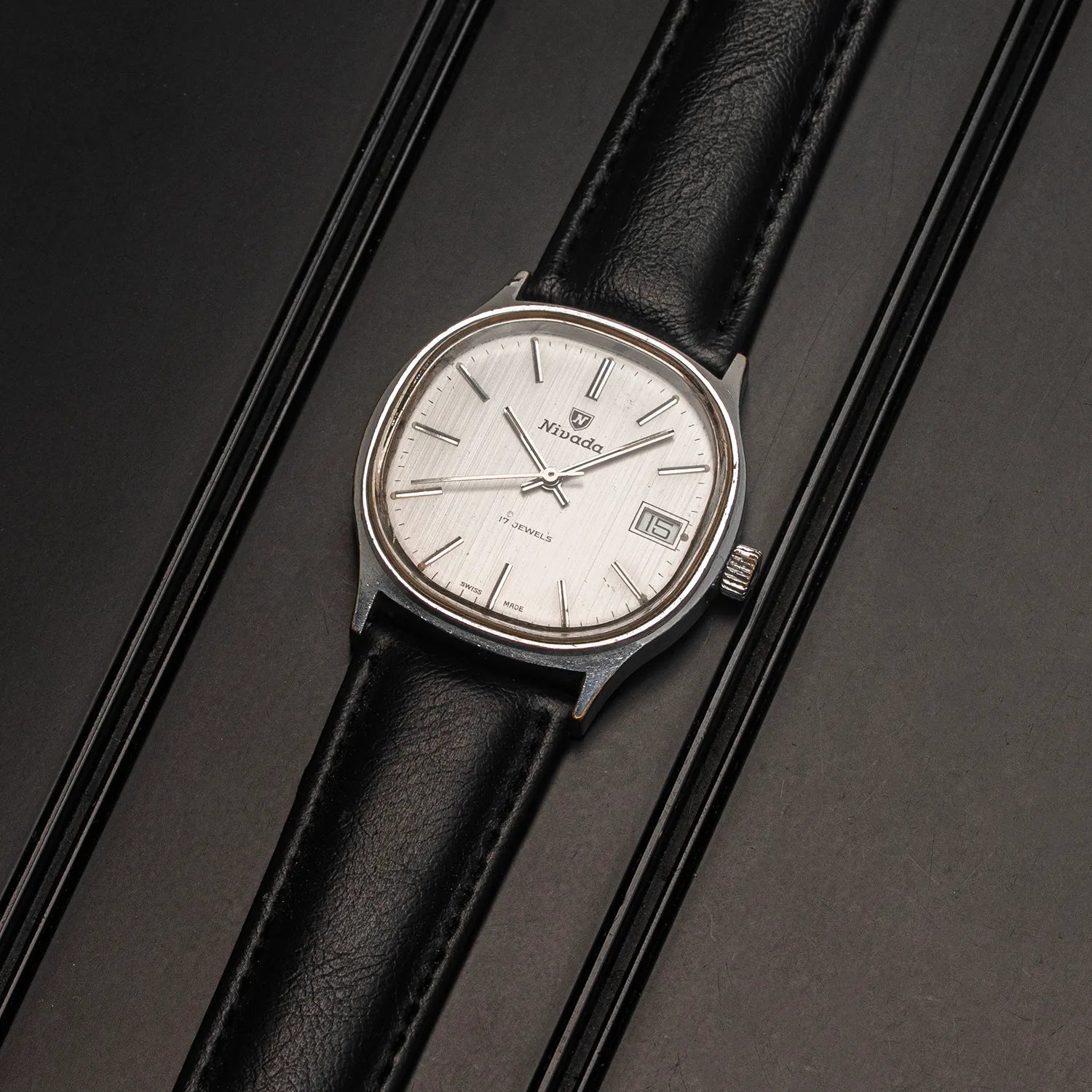 A vintage-style wristwatch Nivada Square with a silver-toned oval case, white dial, and black leather strap, photographed diagonally on a dark textured surface.