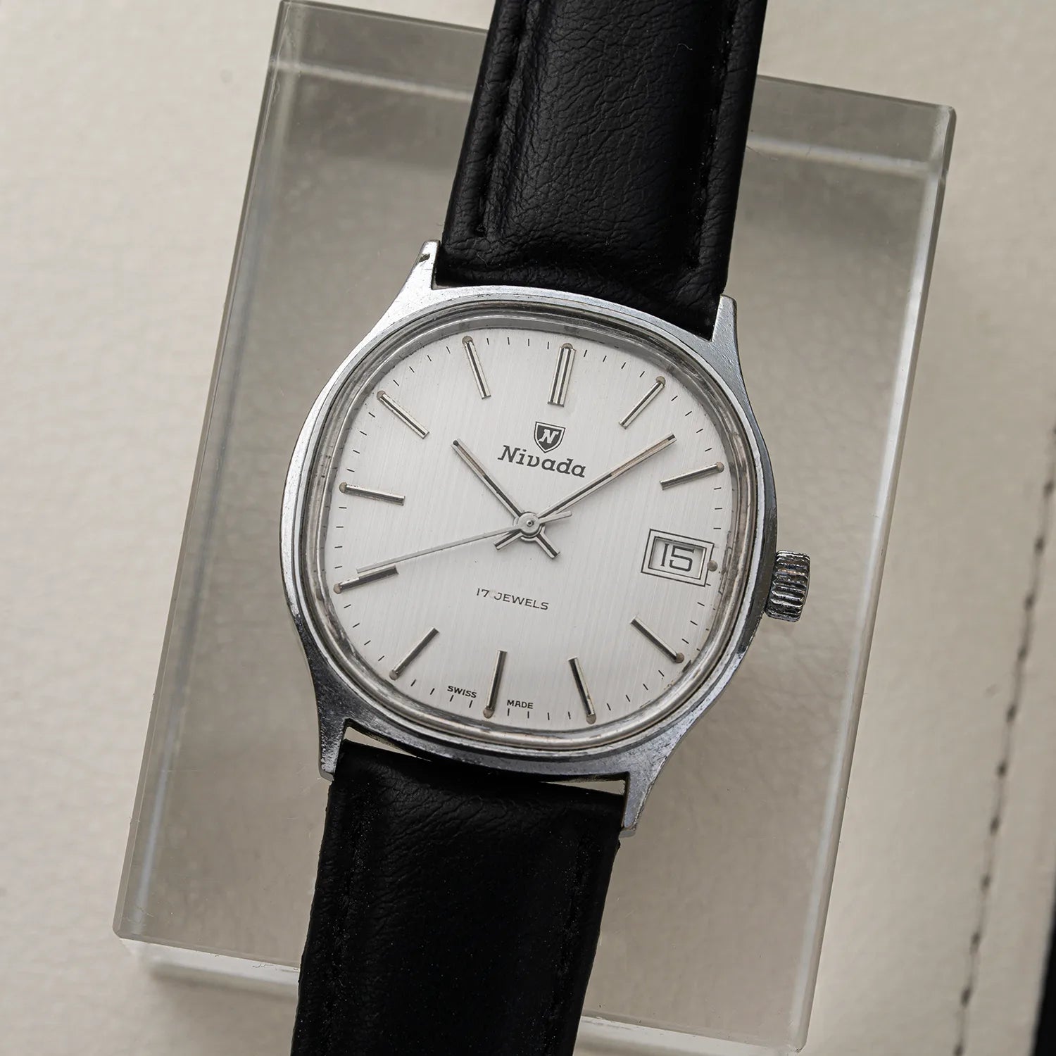 A vintage Niwada watch with a silver-toned metal case, white dial, and black leather strap, displayed on a clear acrylic stand.