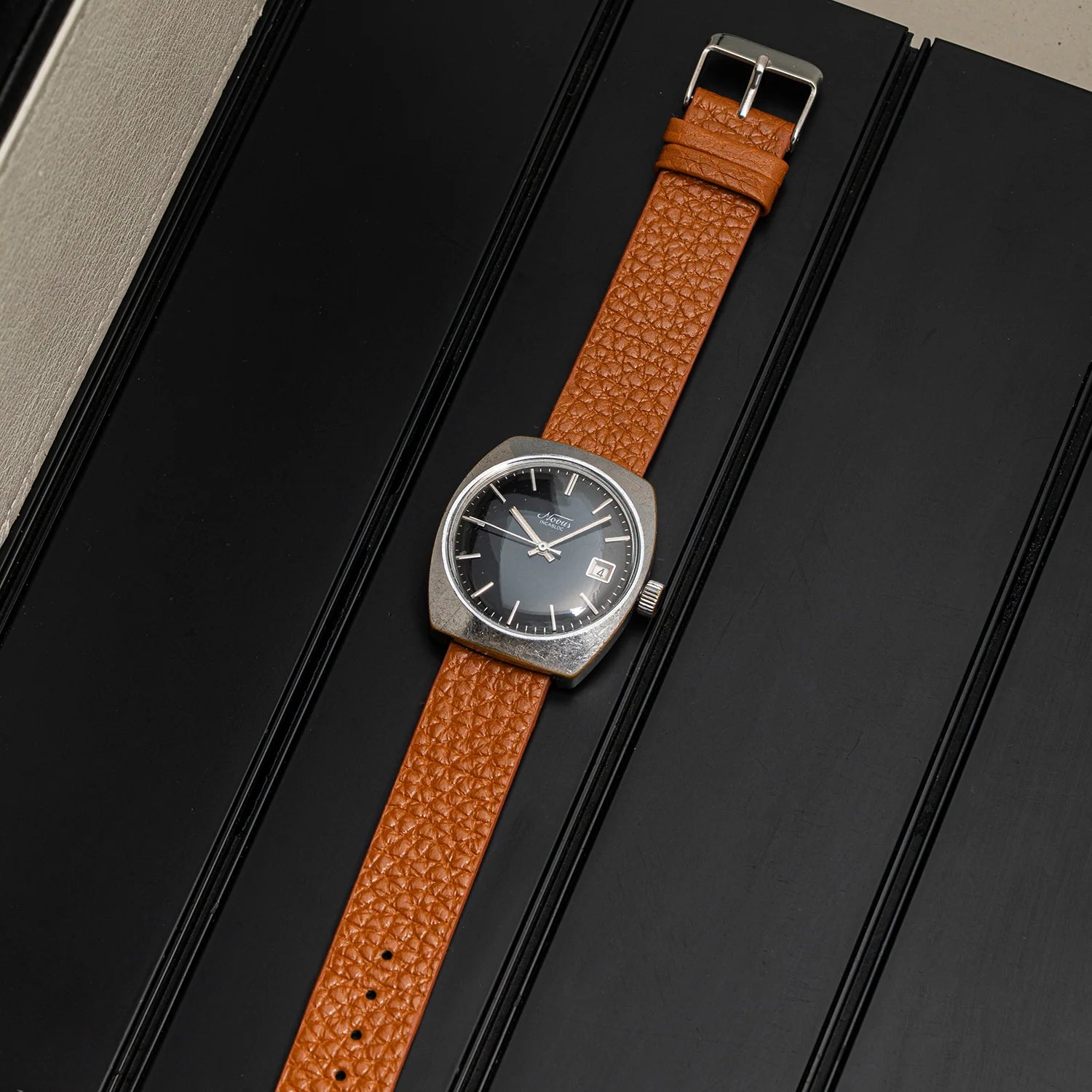 A vintage-style wristwatch Novus, Cal. FE 3612 with a textured brown leather strap and a dark gray dial, displayed diagonally on a black ribbed surface.