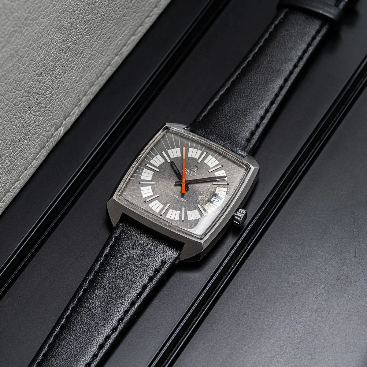 A rectangular silver-tone wristwatch Omega Seamaster, Ref. 166.042, Cal. 565, Men’s Automatic with a black leather strap, shown resting on a dark textured surface next to a light gray textured material. The watch face features a silver dial with white Roman numerals and a single orange second hand.