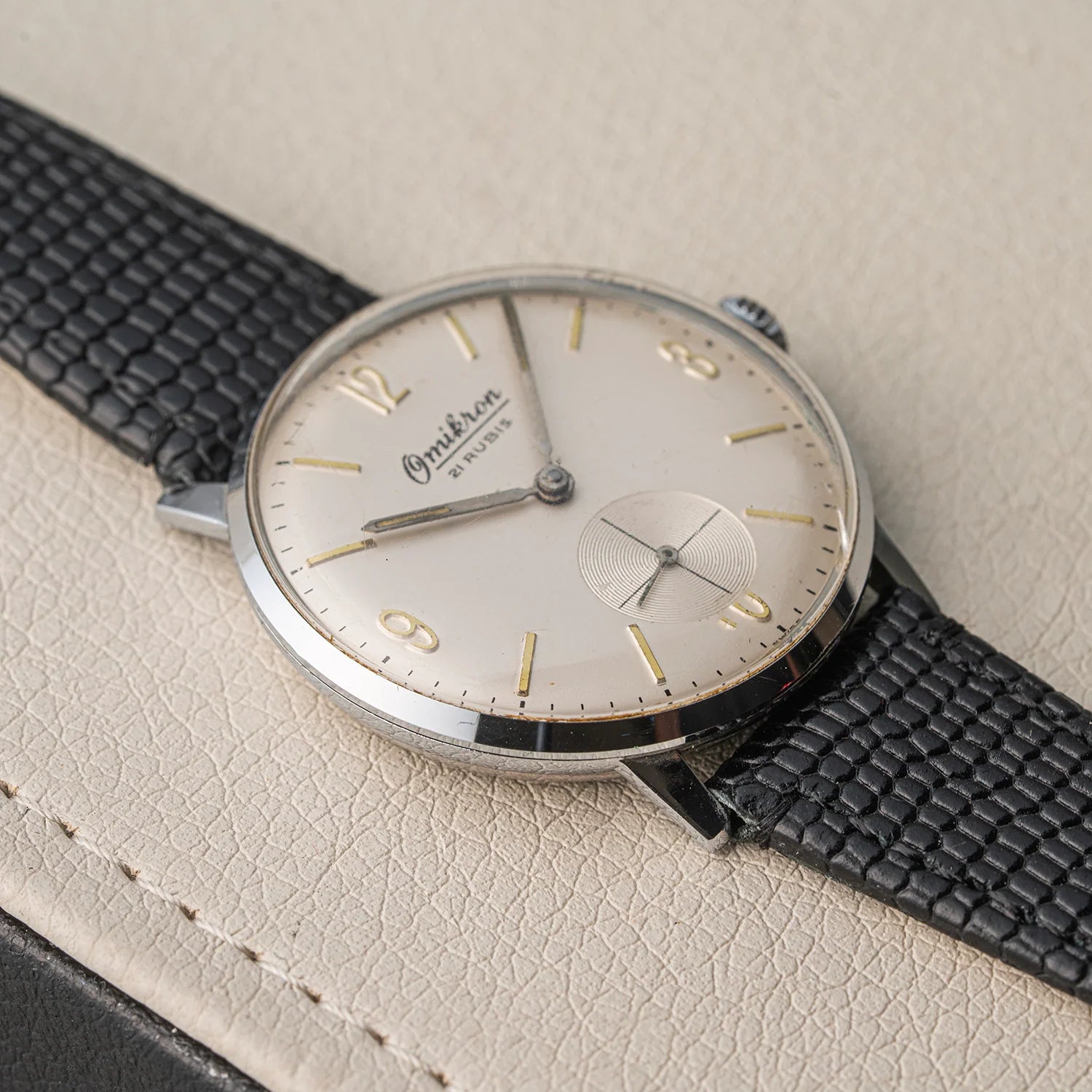 A vintage Omikron, Ref. 3360, Cal. AS 1130 watch with a silver-toned oval case, cream-colored dial, gold-tone numerals and hands, and a textured black leather strap, resting on a light beige textured leather surface.