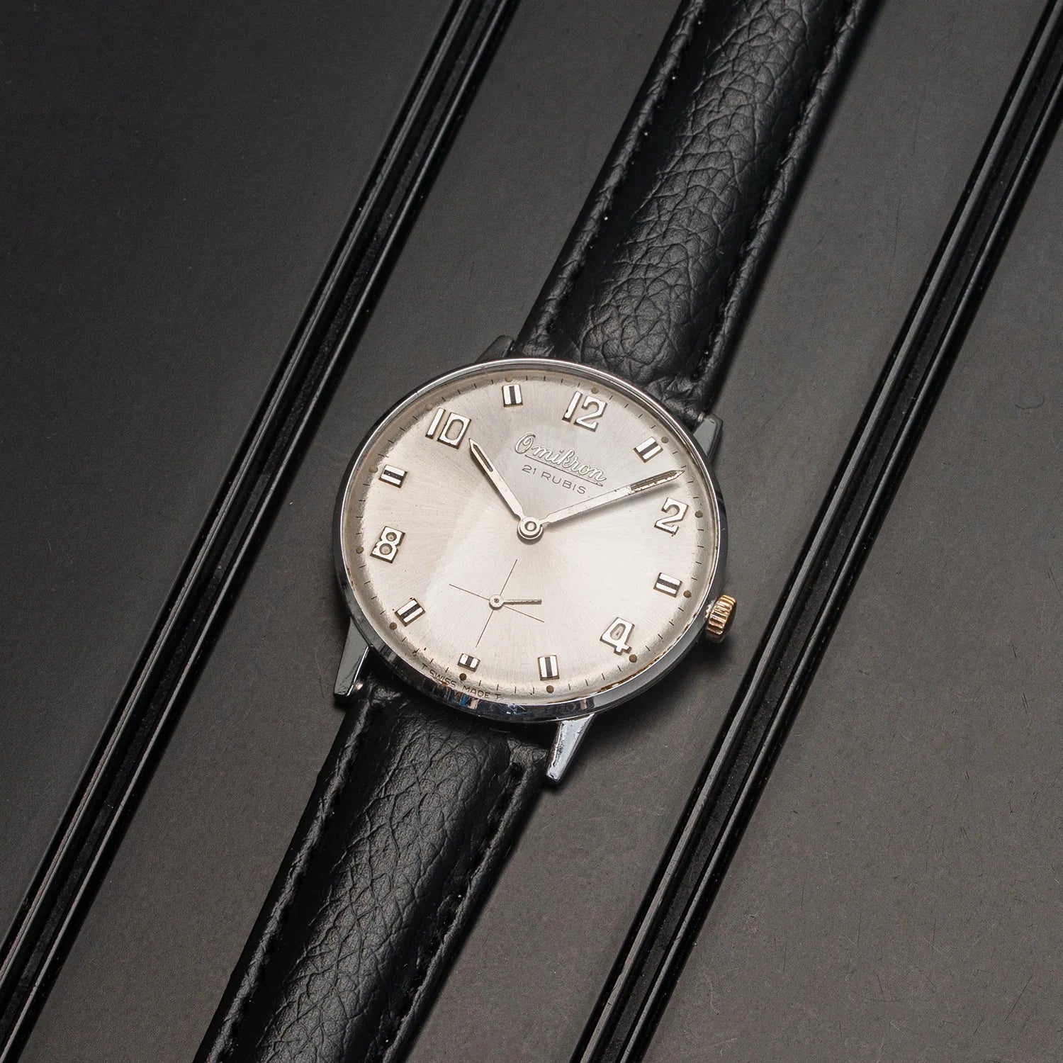 A vintage-style wristwatch Omikron AS 1130 with a silver-toned oval dial, black leather strap, and gold-tone crown, displayed diagonally on a dark textured background.