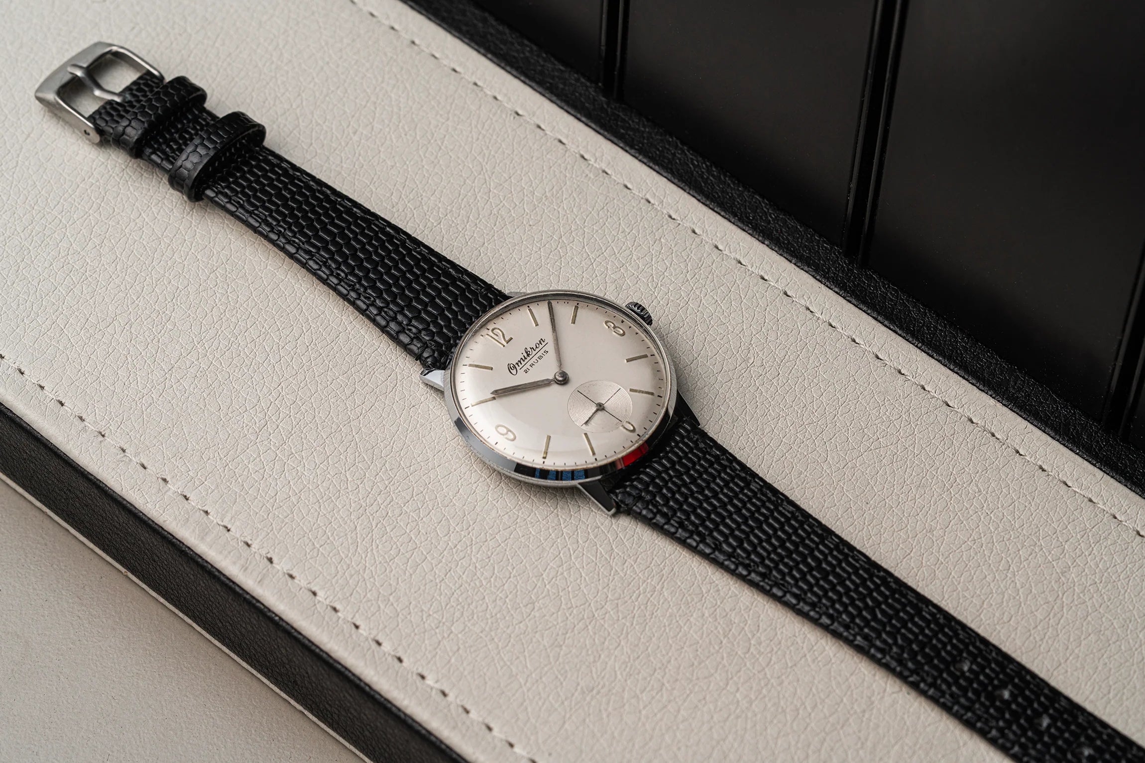 A vintage-style wristwatch Omikron, Ref. 3360, Cal. AS 1130 with a silver-toned round dial, black textured strap, and a small red, white, and blue accent on the lower dial, resting diagonally on a light beige leather surface.