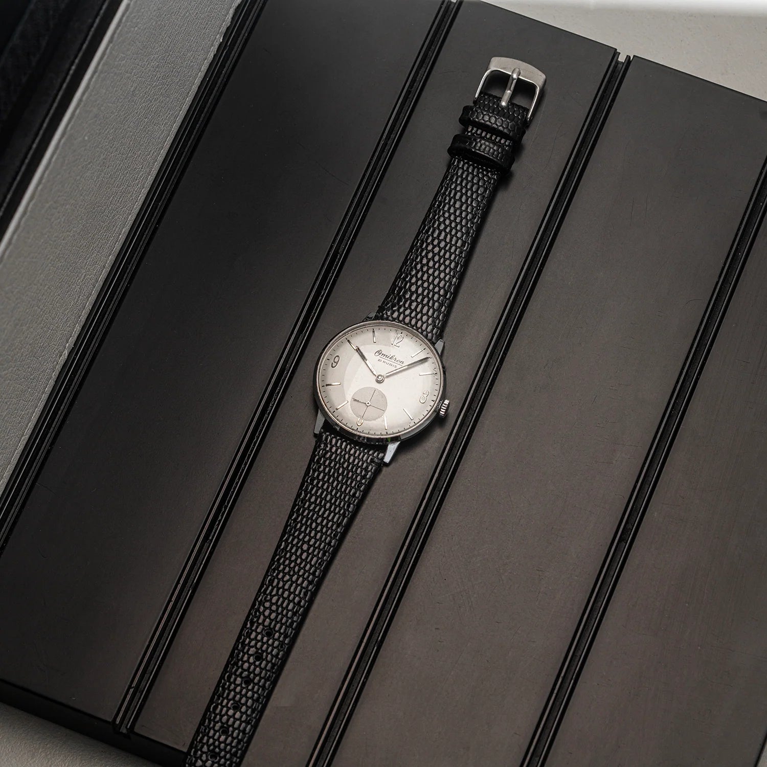 A wristwatch Omikron, Ref. 3360, Cal. AS 1130 with a black textured mesh strap and a silver-toned round dial, displayed diagonally on a dark gray presentation box.