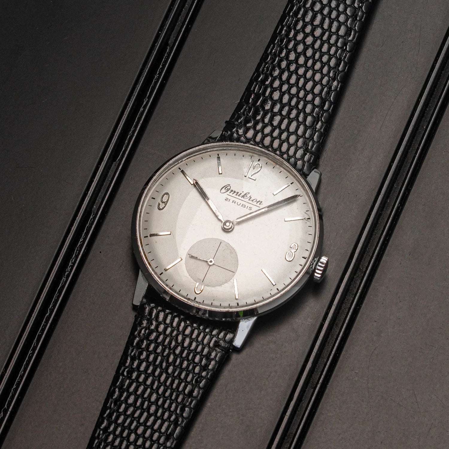 A vintage Omikron, Ref. 3360, Cal. AS 1130 watch with a silver dial and a textured black strap, presented diagonally on a dark textured background.