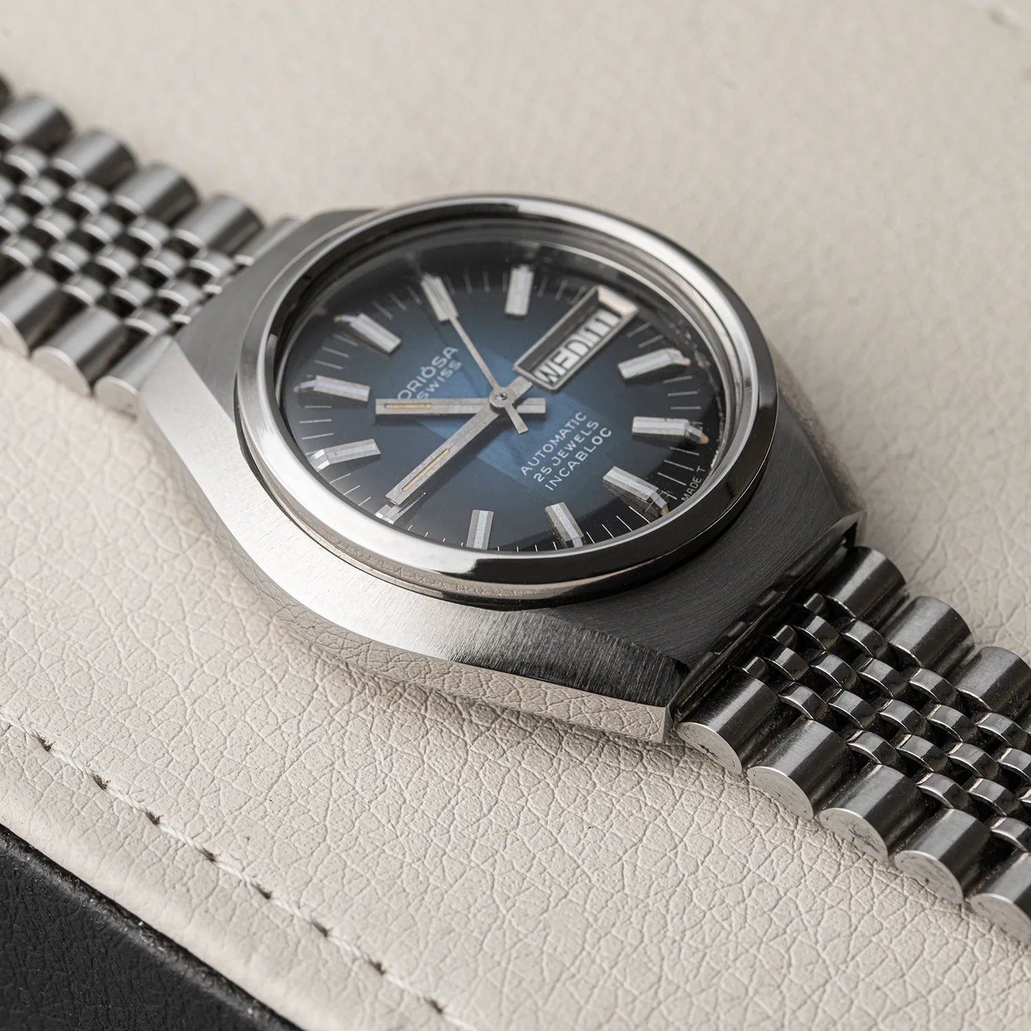 A close-up of a wristwatch Oriosa, ETA 2789 with a silver-toned metal case and bracelet, featuring a dark blue dial with silver hour markers and hands, resting on a textured light beige surface.