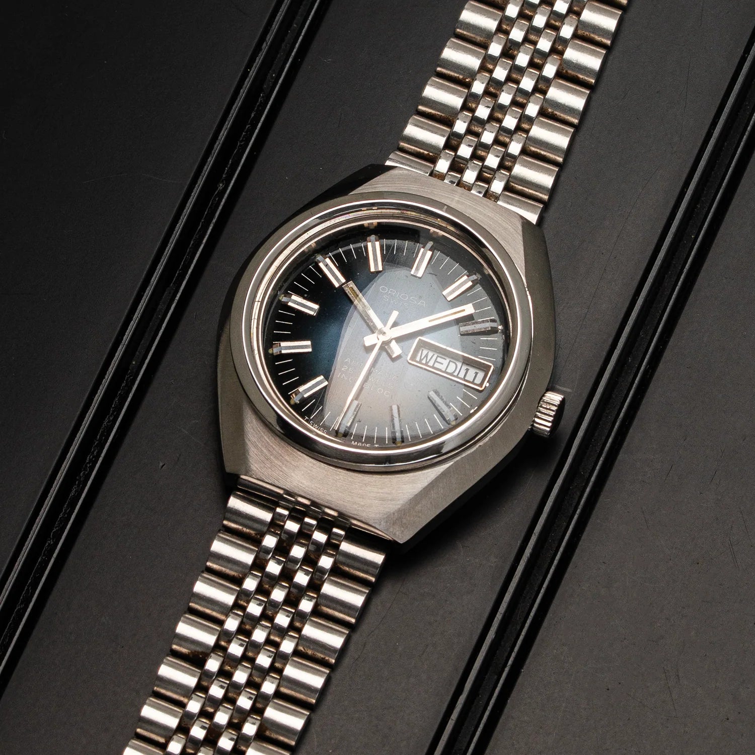 A silver-toned metal wristwatch with a matching link bracelet, featuring a blue-gray sunburst dial, silver hour markers, and a day-date window at 3 o'clock, displayed on a dark textured background.