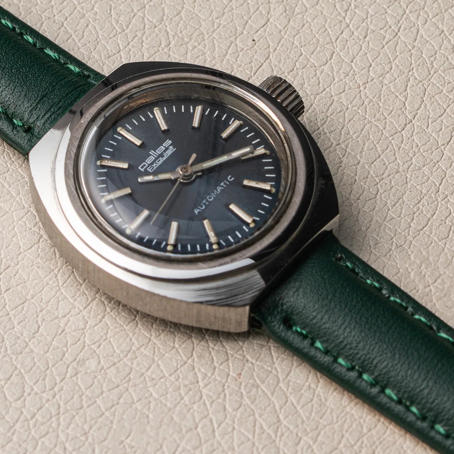 A vintage-style wristwatch Pallas Exquisit ETA 2670 with a dark blue dial, silver-toned metal case, and a dark green leather strap with visible stitching, photographed on a textured light beige surface.