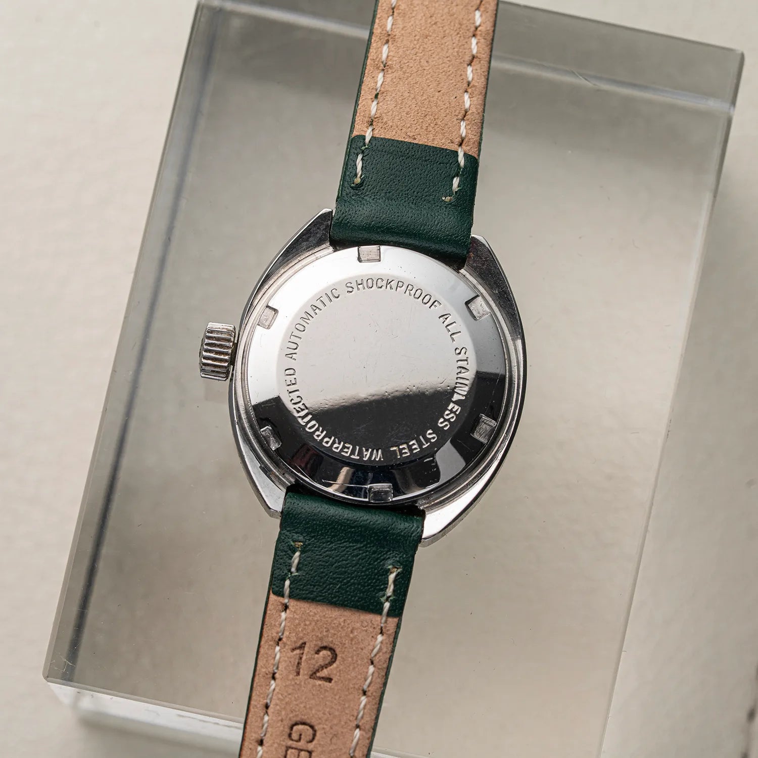 Back view of a wristwatch Pallas Exquisit ETA 2670 with a stainless steel case and a green leather strap with tan stitching, displayed on a clear acrylic stand against a light beige background.
