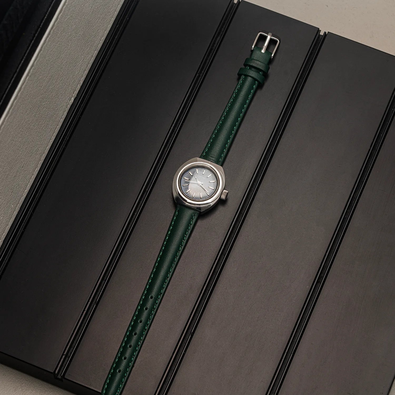 A wristwatch Pallas Exquisit ETA 2670 with a dark green leather strap and a silver-toned round case, displayed diagonally on a dark textured surface.
