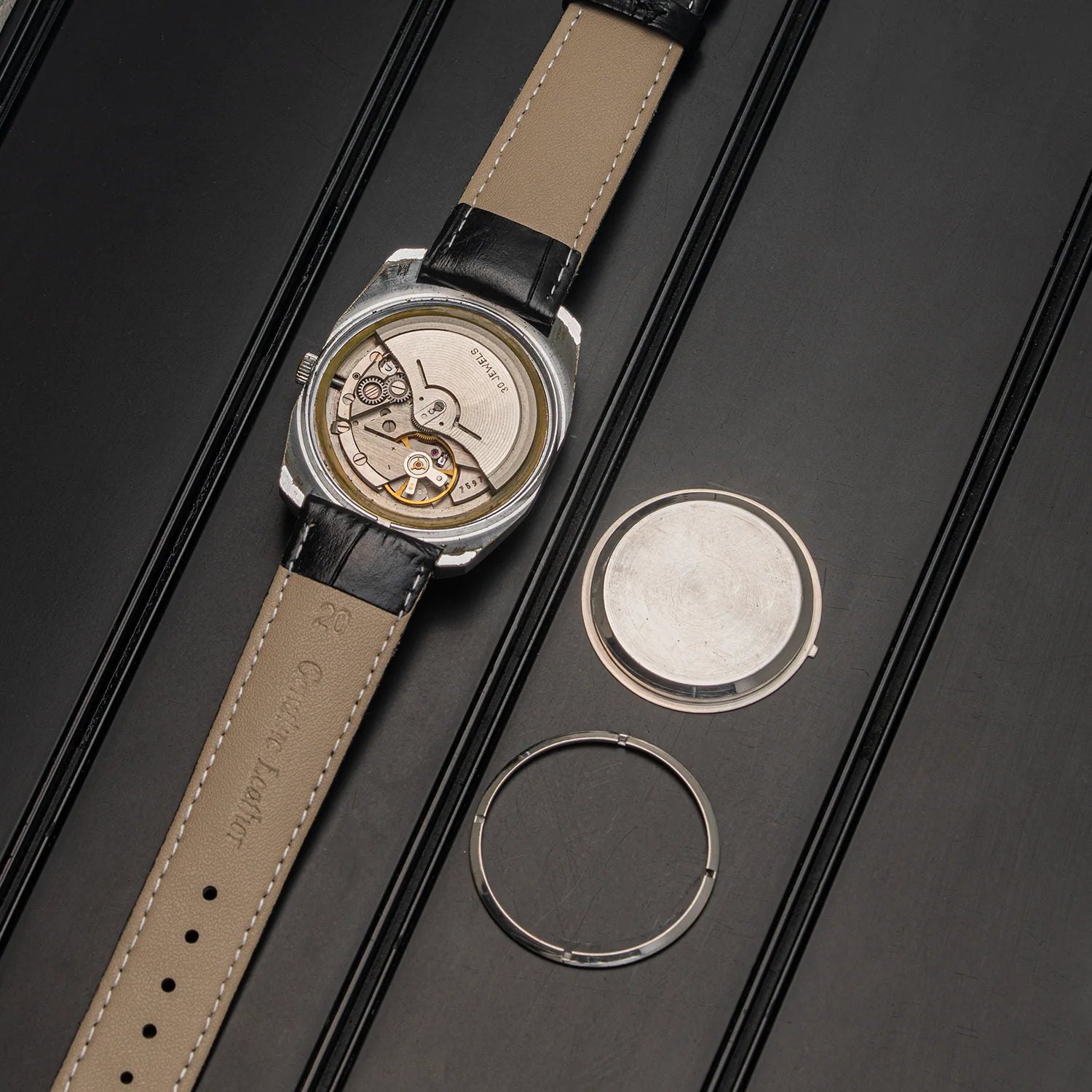 A luxury wristwatch with a silver-toned case, a beige leather strap, and a visible mechanical movement, displayed diagonally on a dark textured background alongside its circular metal components.