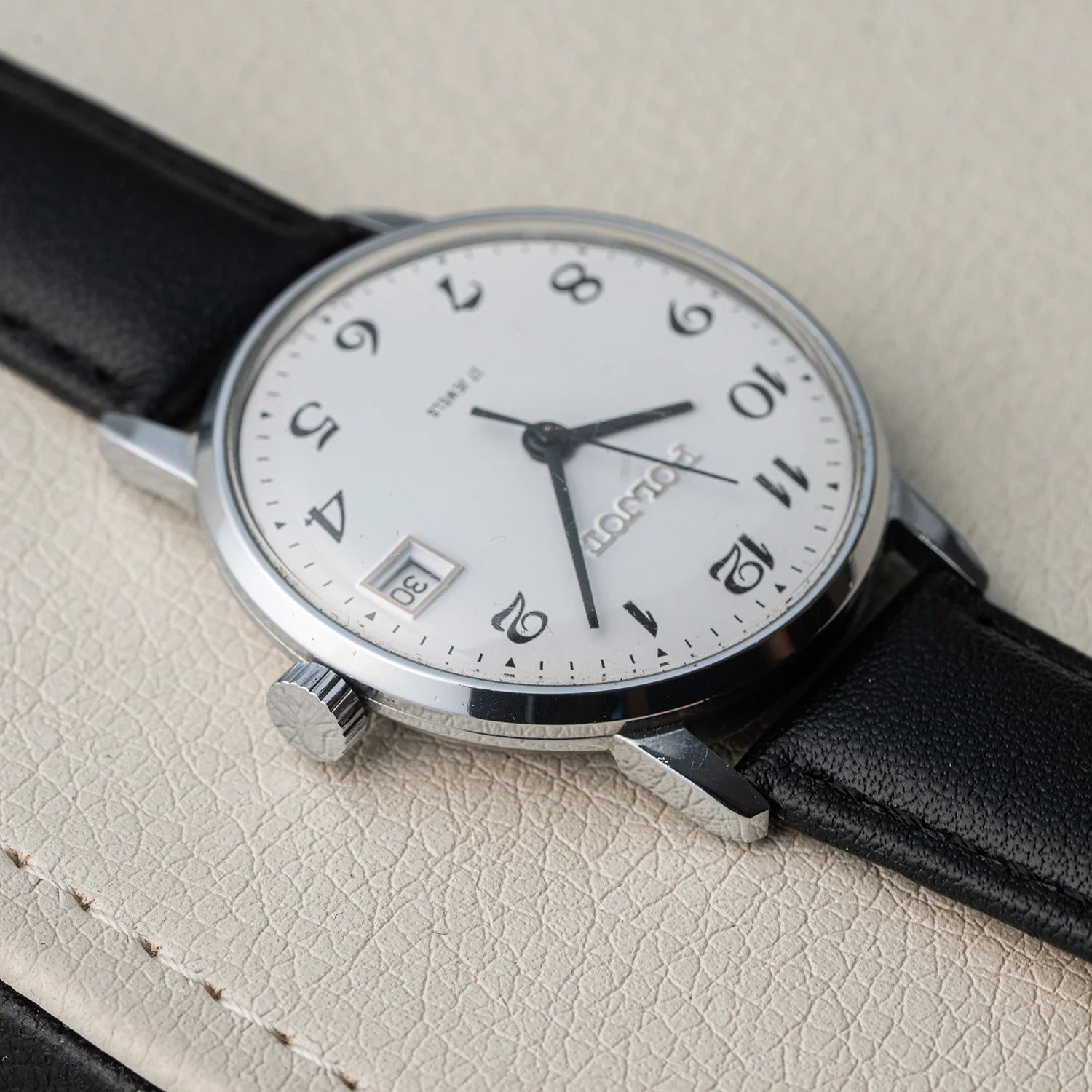 A close-up of a Longines wristwatch Poljot 2614.2H with a silver-toned case, white dial, black Arabic numerals, date window, and black leather strap, resting on a textured light beige surface.