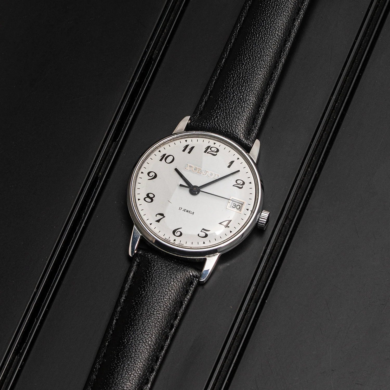 A vintage-style wristwatch Poljot 2614.2H with a silver-toned oval case, white dial, black numerals and hands, and a black leather strap, photographed on a dark textured background.