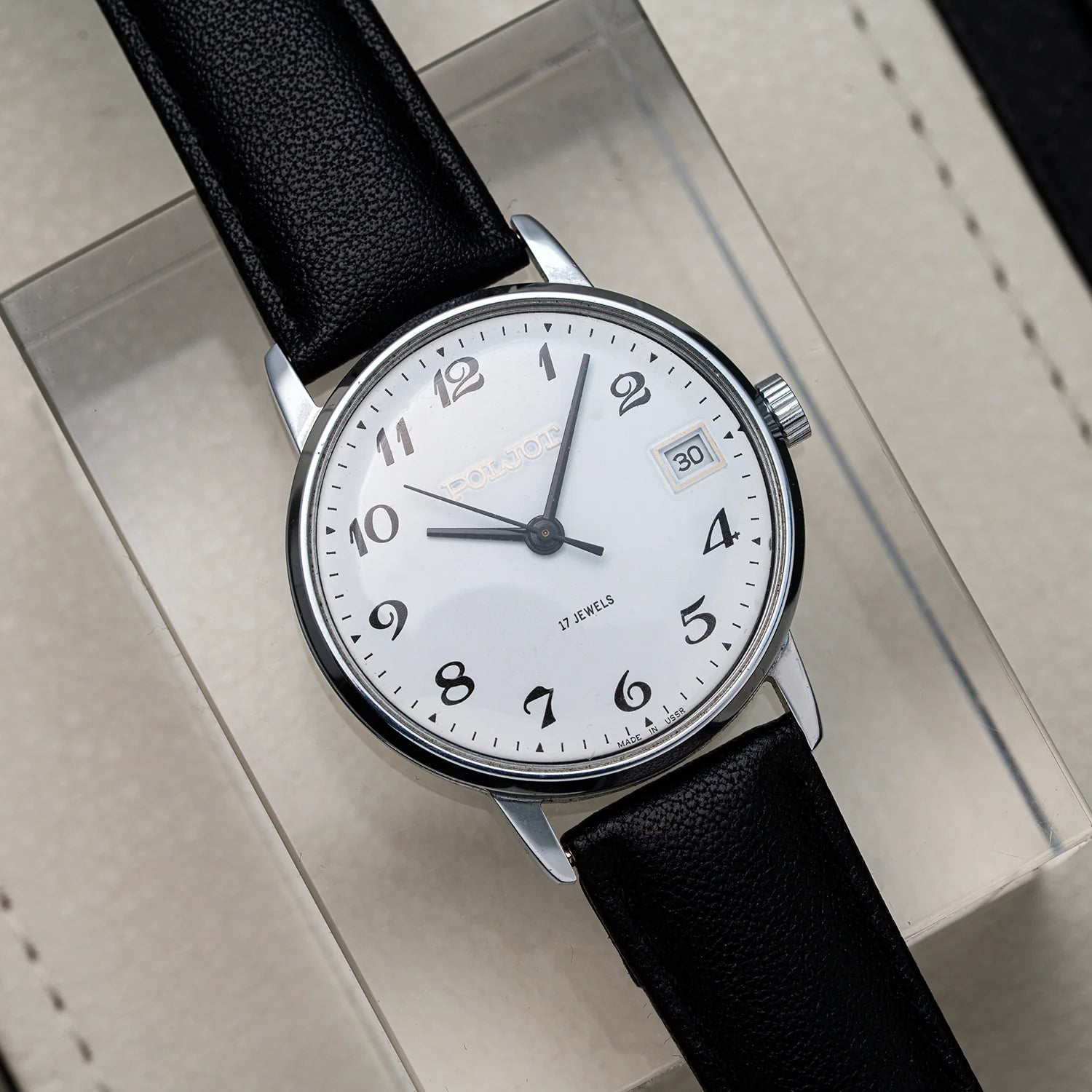 A wristwatch Poljot 2614.2H with a black leather strap, silver-toned case, and a white dial featuring black Arabic numerals and a date window, displayed on a light beige presentation box.