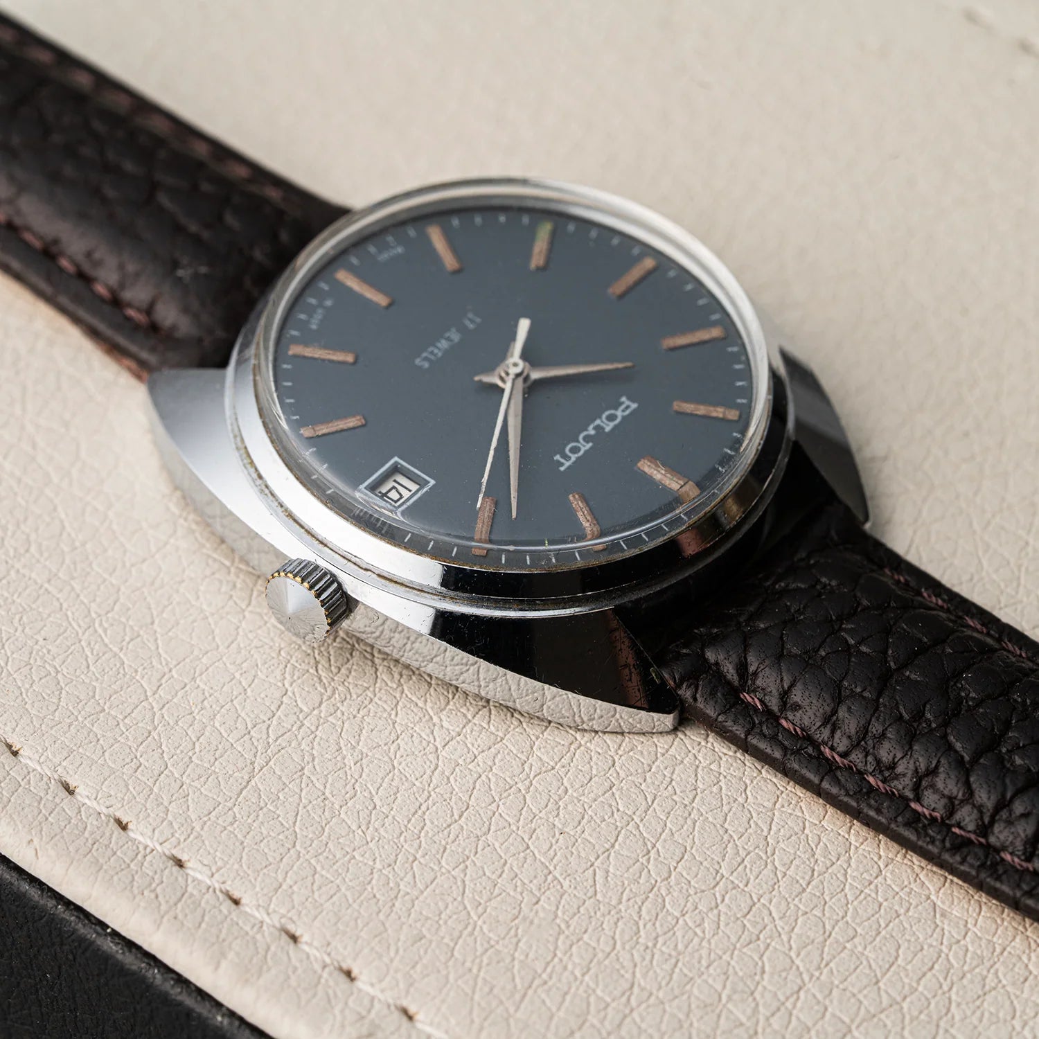 A vintage-style wristwatch Poljot, Cal. 2614.2H with a dark gray dial, rose gold-toned hour markers and hands, and a silver-tone case, resting on a textured cream-colored leather surface.