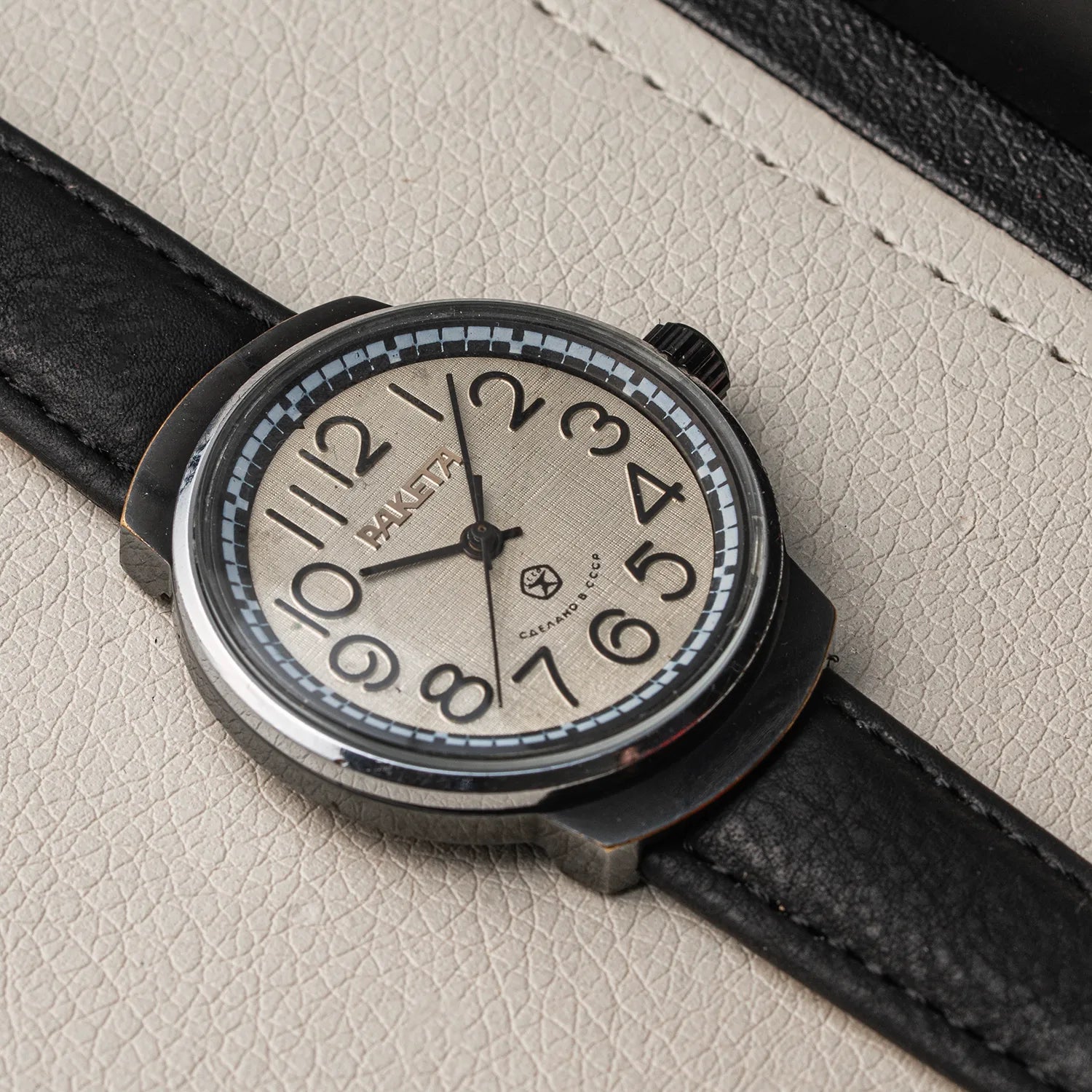 A close-up of a Raketa Big Numerals wristwatch with a black leather strap and a beige dial, resting on a textured light beige surface.