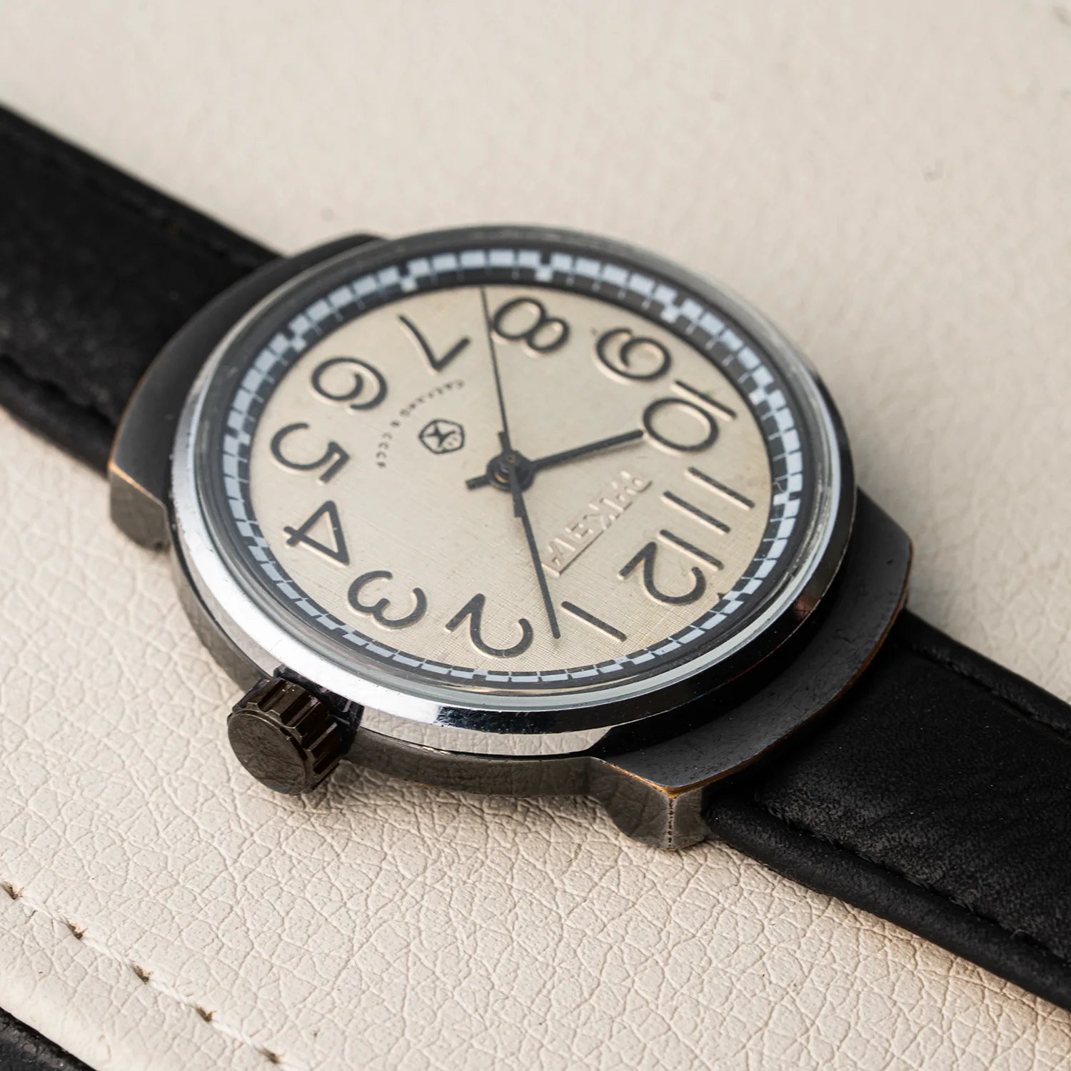A close-up of a vintage-style wristwatch Raketa Big Numeralswith a black leather strap, black case, and a cream-colored dial featuring bold black Arabic numerals. The brand name 'TITAN' is visible on the dial.