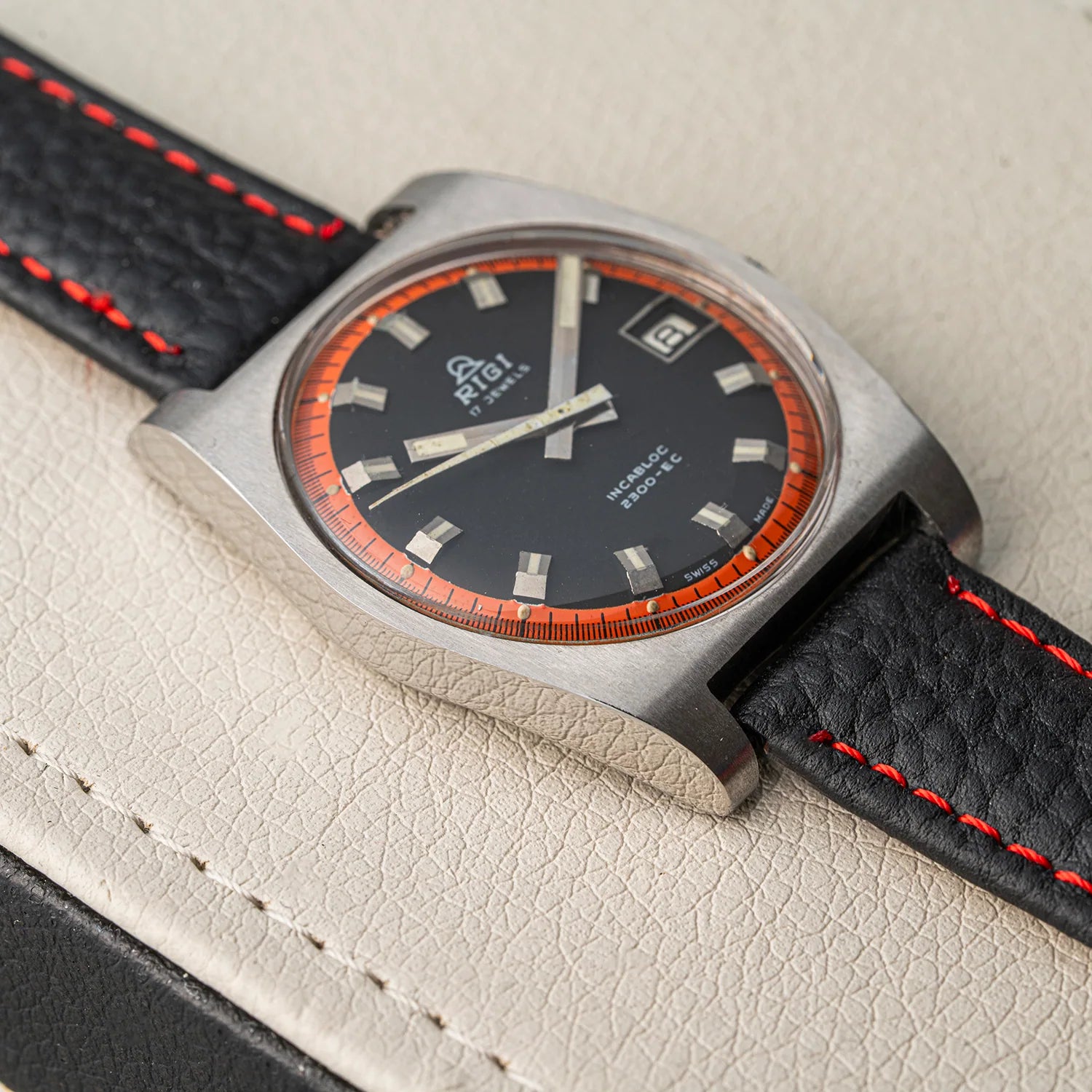 A vintage-style wristwatch Rigi 2300-EC with a square brushed-metal case, black dial, orange ring around the bezel, and a black leather strap with red stitching, photographed close up on a light textured surface.