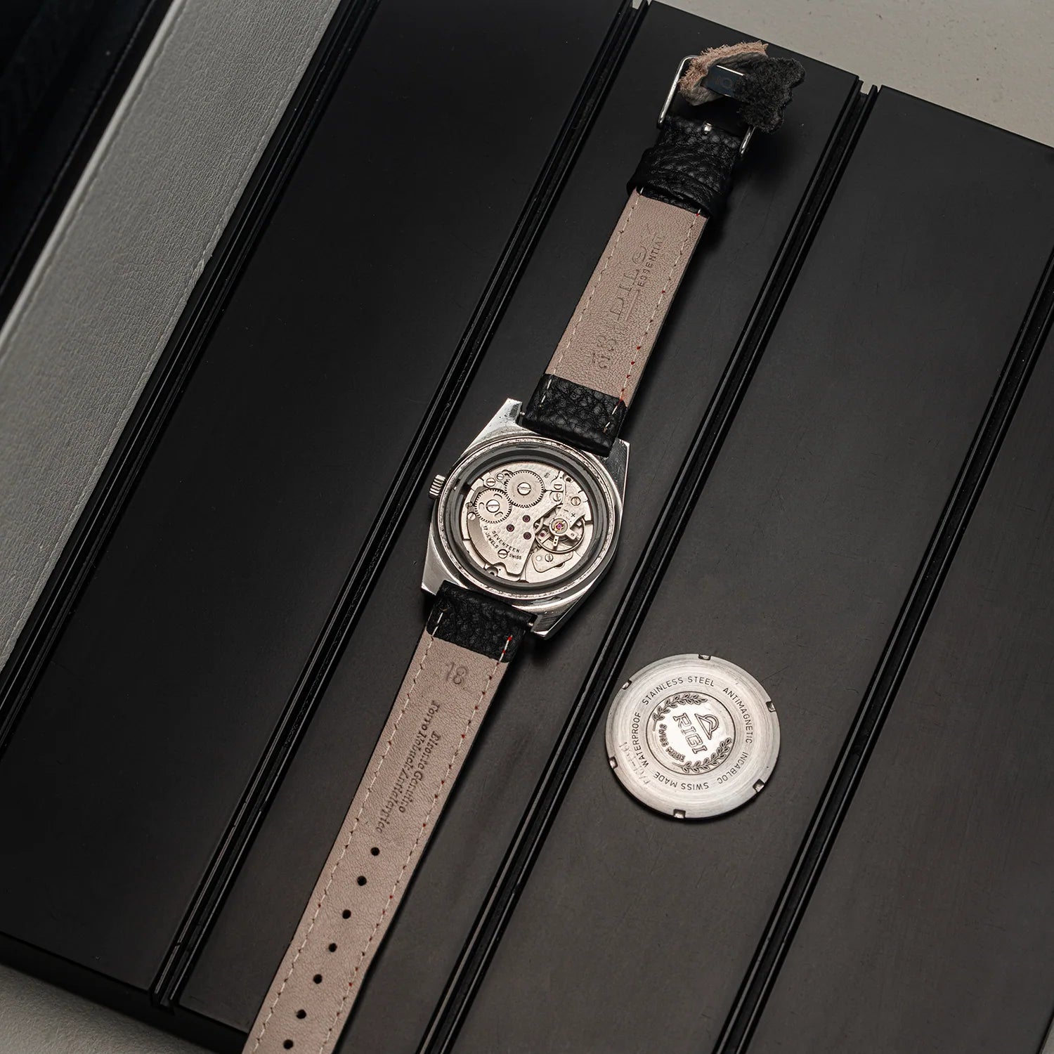 A vintage-style Rigi 2300-EC watch with a silver-toned case and a beige leather strap, displayed diagonally on a black presentation box next to its circular backplate.